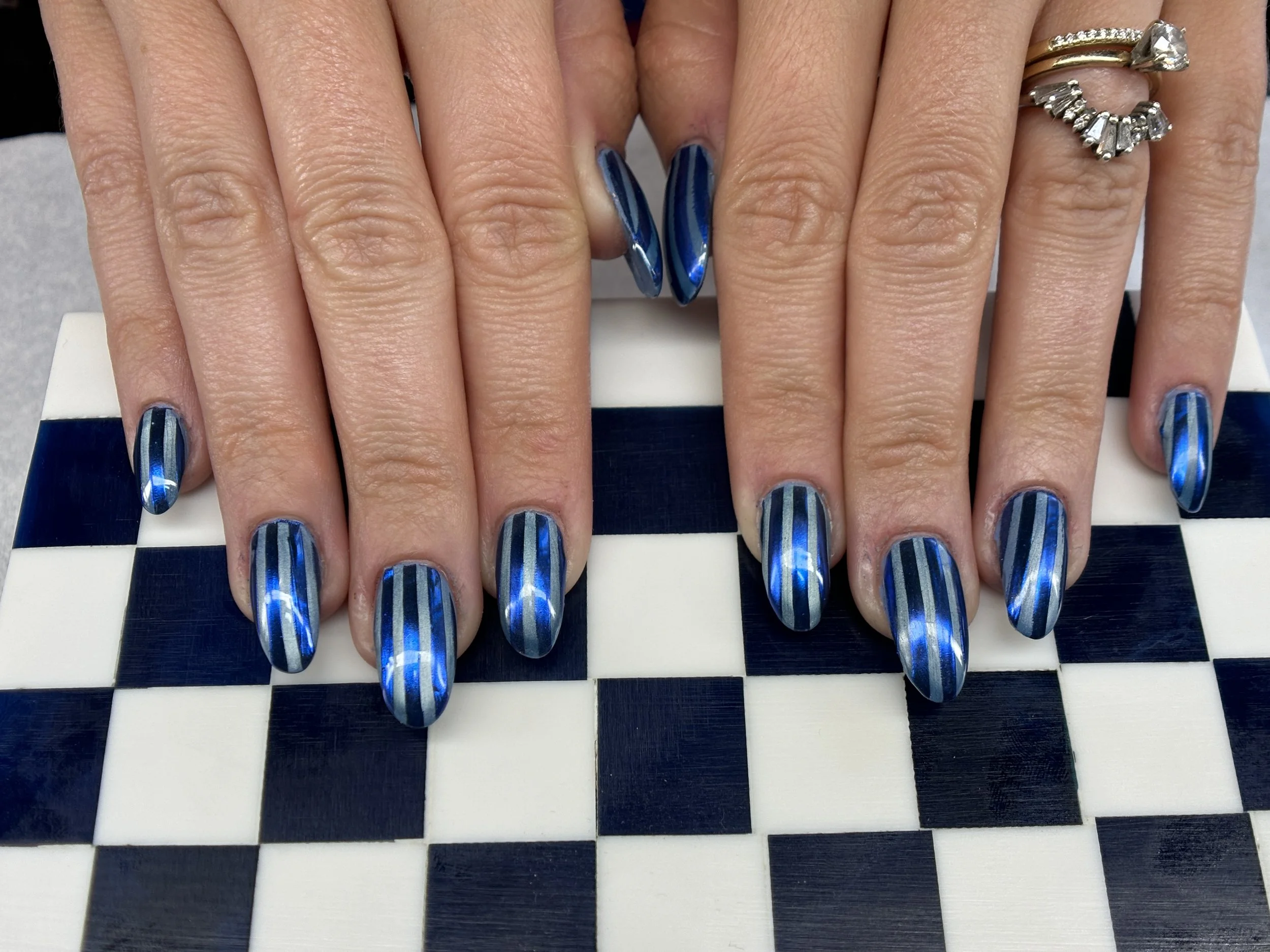 Close-up of a person's hand with blue striped long nails, displaying multiple rings on the fingers, resting on a black and white checkered surface.