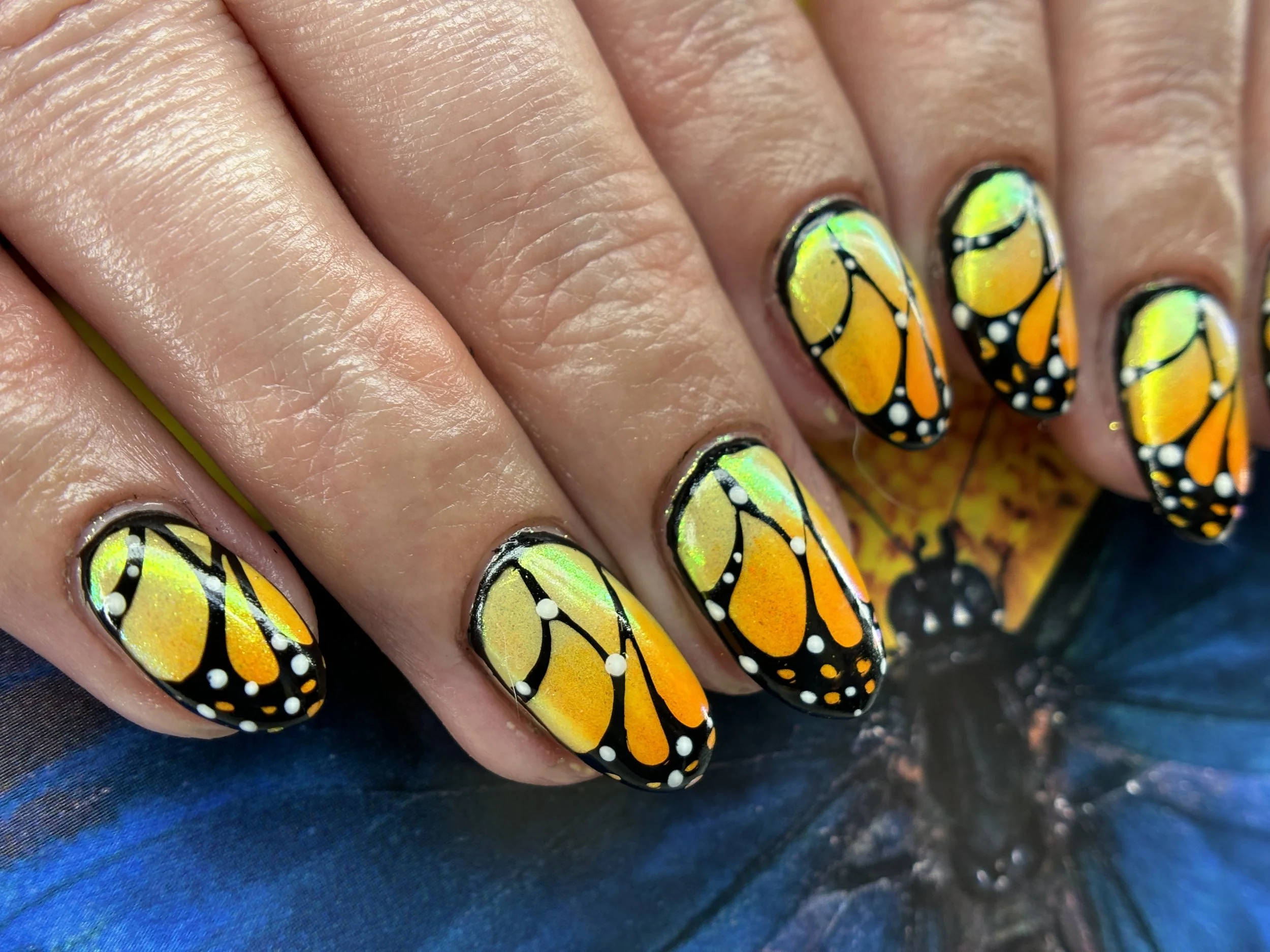 Close-up of a hand with butterfly-themed nail art on fingernails, featuring colorful orange, yellow, green, and black designs resembling butterfly wings.