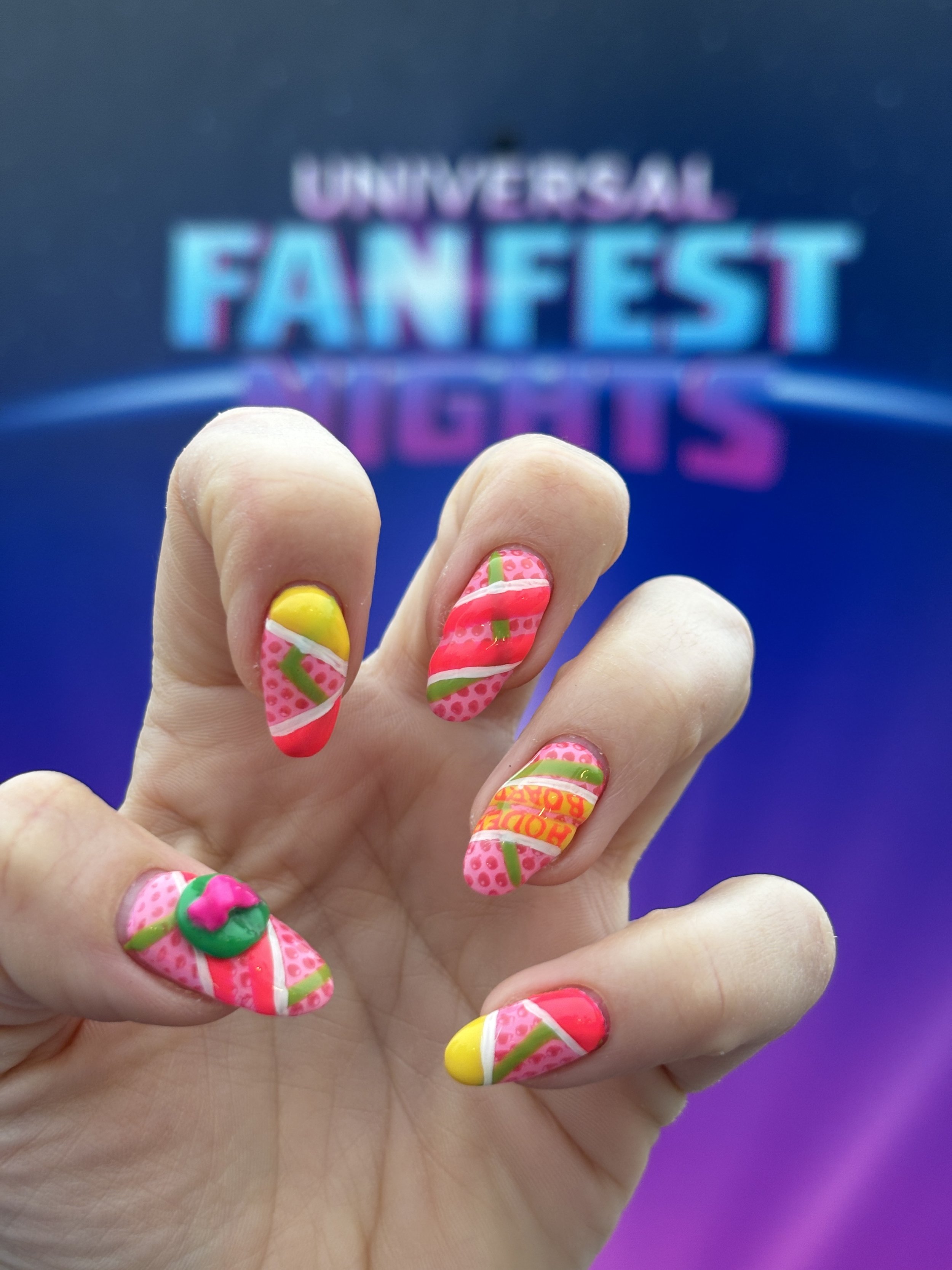 Close-up of a hand with colorful, candy-themed nail art against a blurry blue and purple background with the words 'Universal Fan Fest'