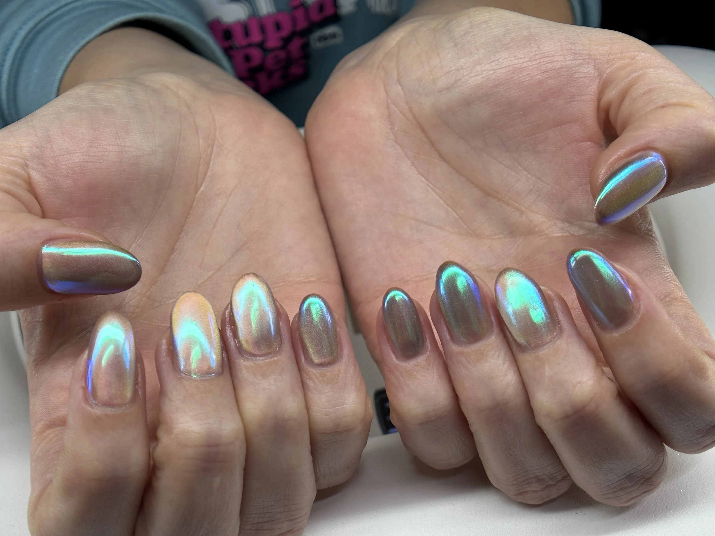 Close-up of a hand showing rainbow-colored, metallic, oval-shaped nails with a glossy finish, held over a surface.