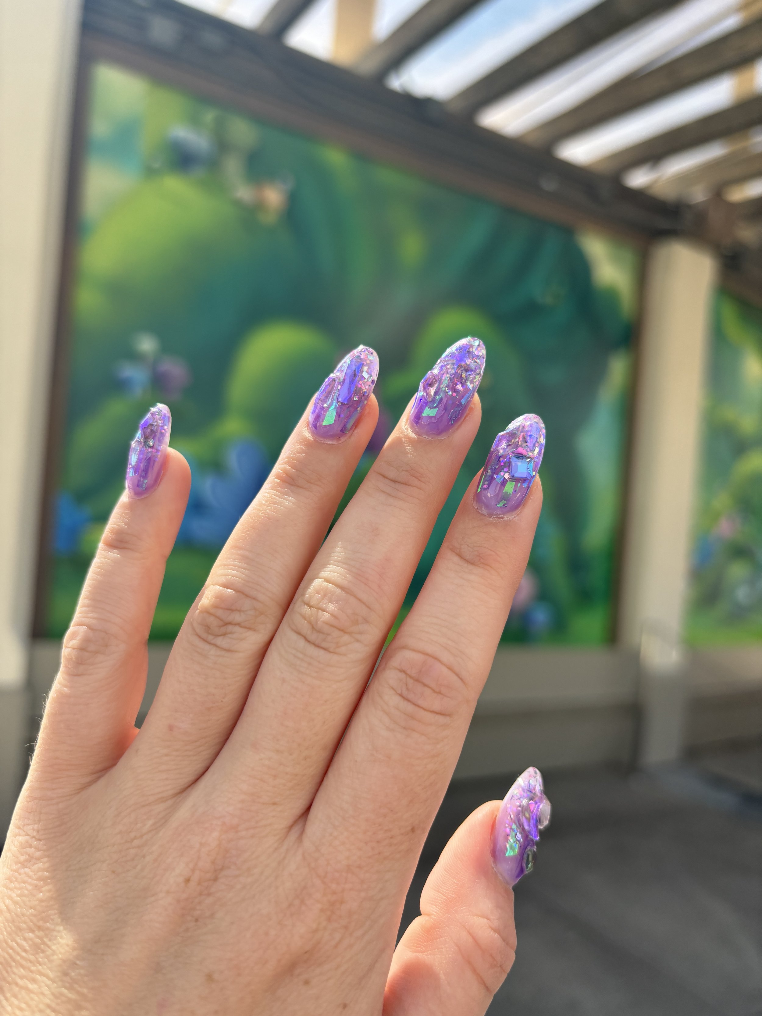 Close-up of a person's hand with purple and iridescent glittery nail polish, fingers slightly splayed, with a blurred outdoor background featuring a structure with wooden beams and a colorful mural or painting.