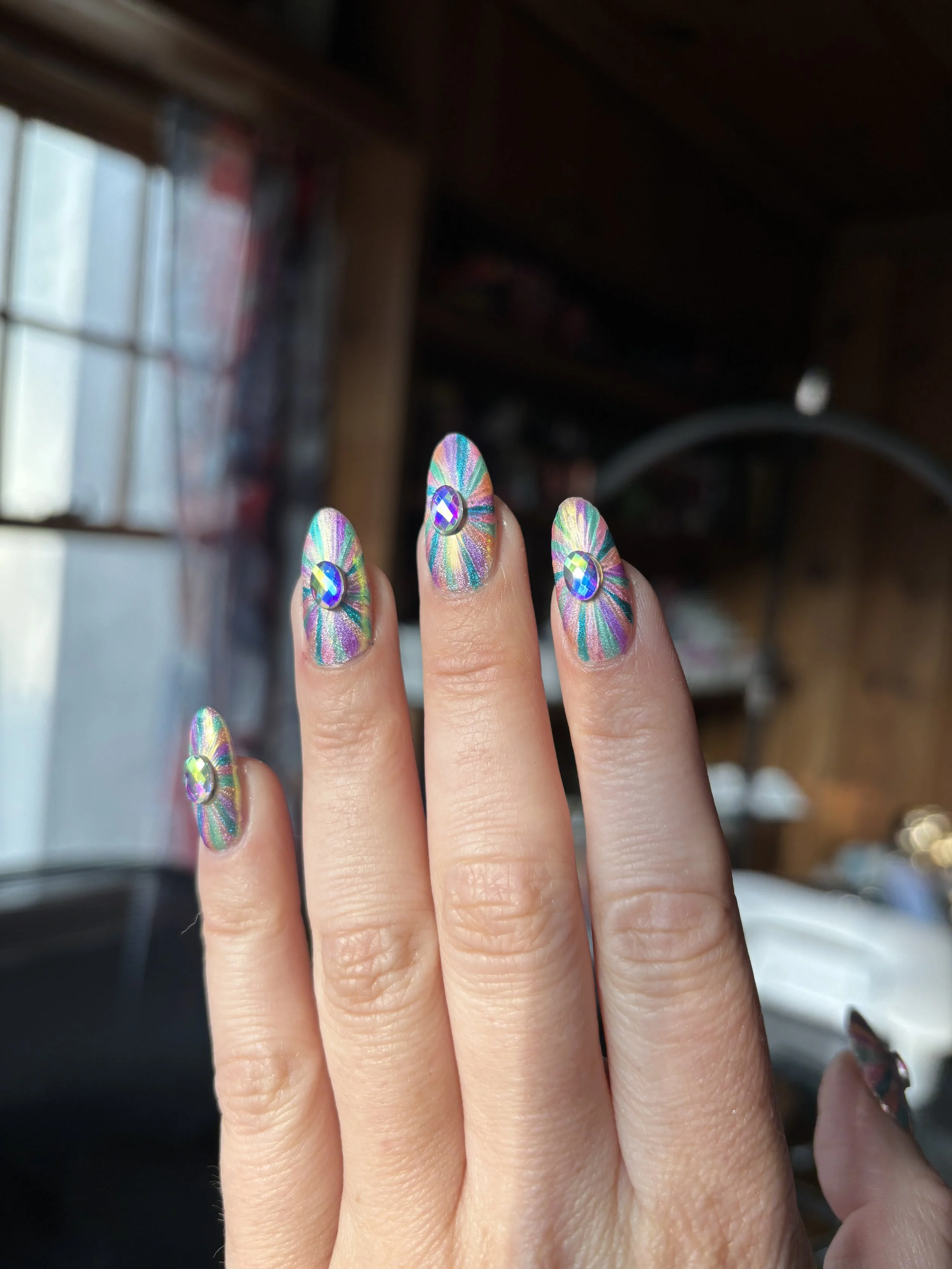 A person's hand showing nails with rainbow-colored, holographic, sunburst nail art, each including a small, shiny gemstone in the center.