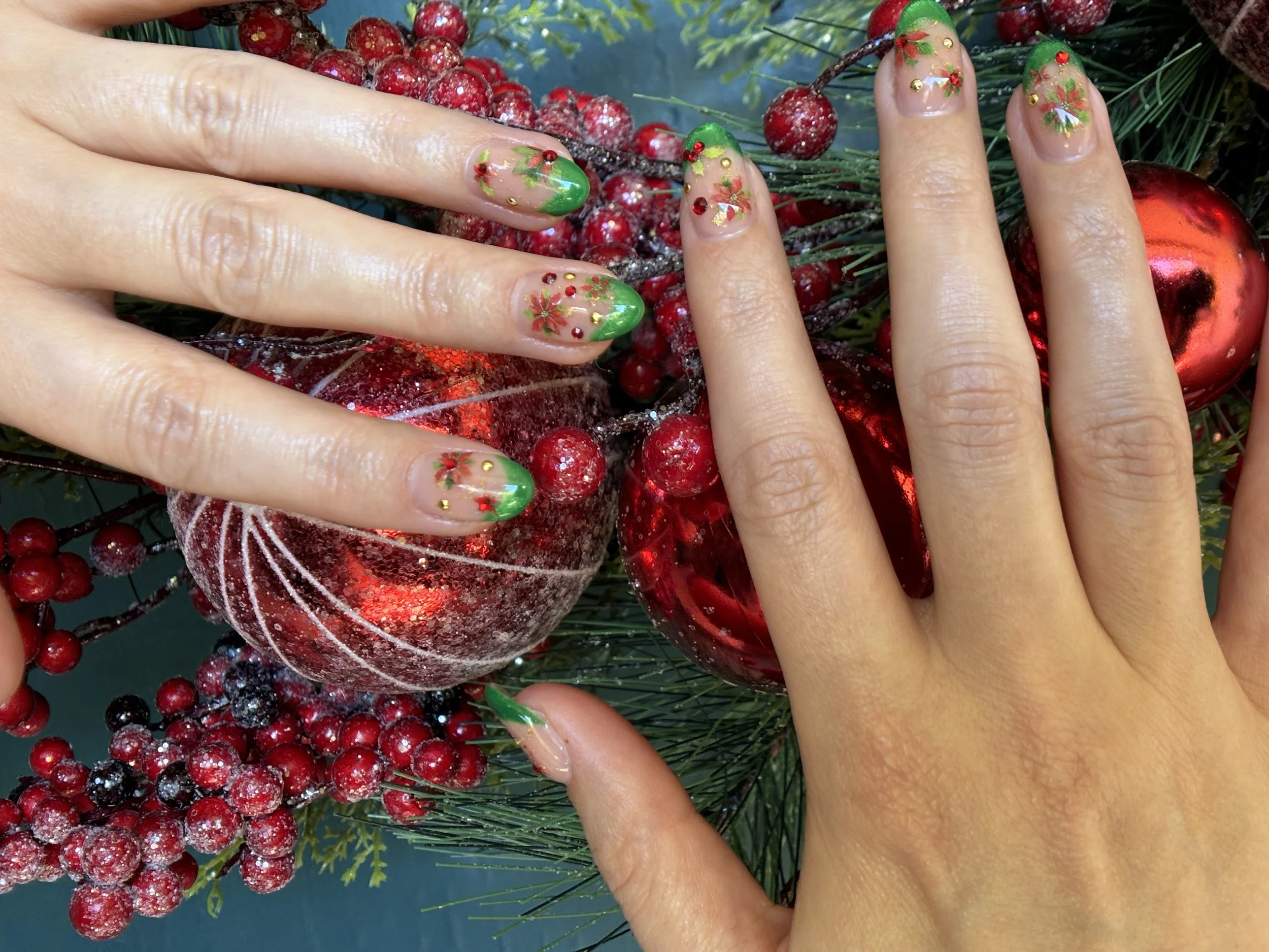 Close-up of hands with decorated Christmas nails placed on a festive arrangement of red berries, green foliage, and red ornaments.