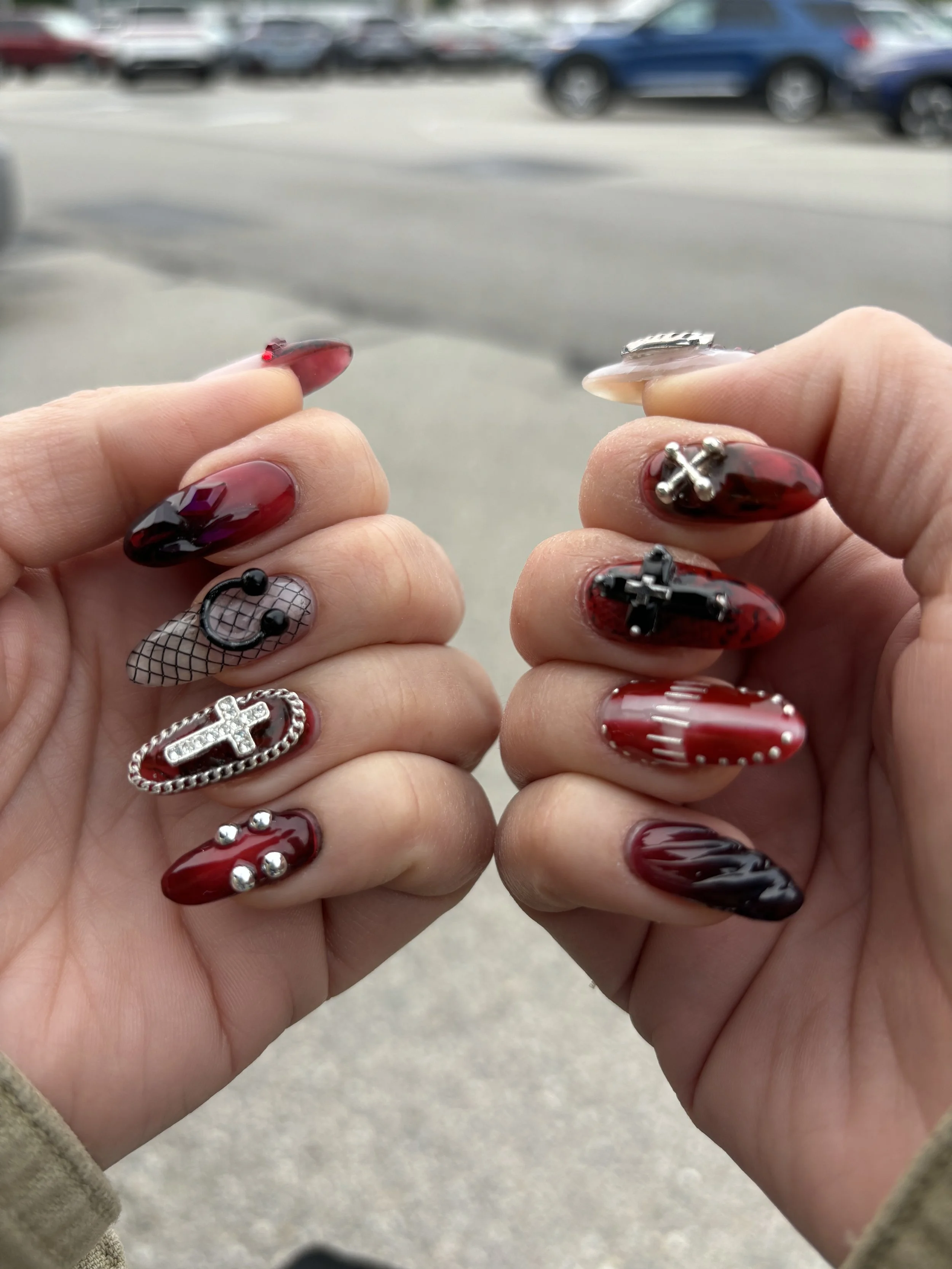 Close-up of hands with decorated nails, showing various intricate nail designs and embellishments, with a parking lot in the background.