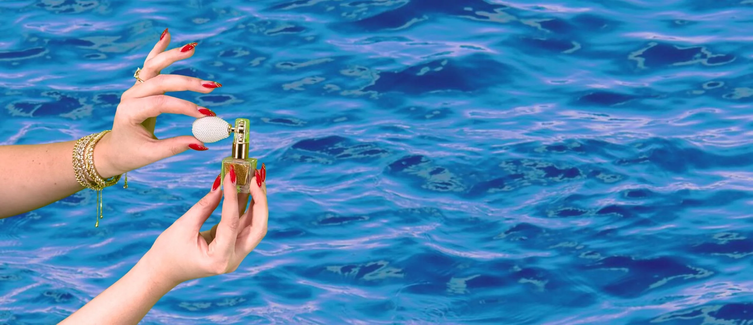A woman's hand with red painted nails and gold jewelry holding a perfume bottle with a spherical cap in front of a body of water.