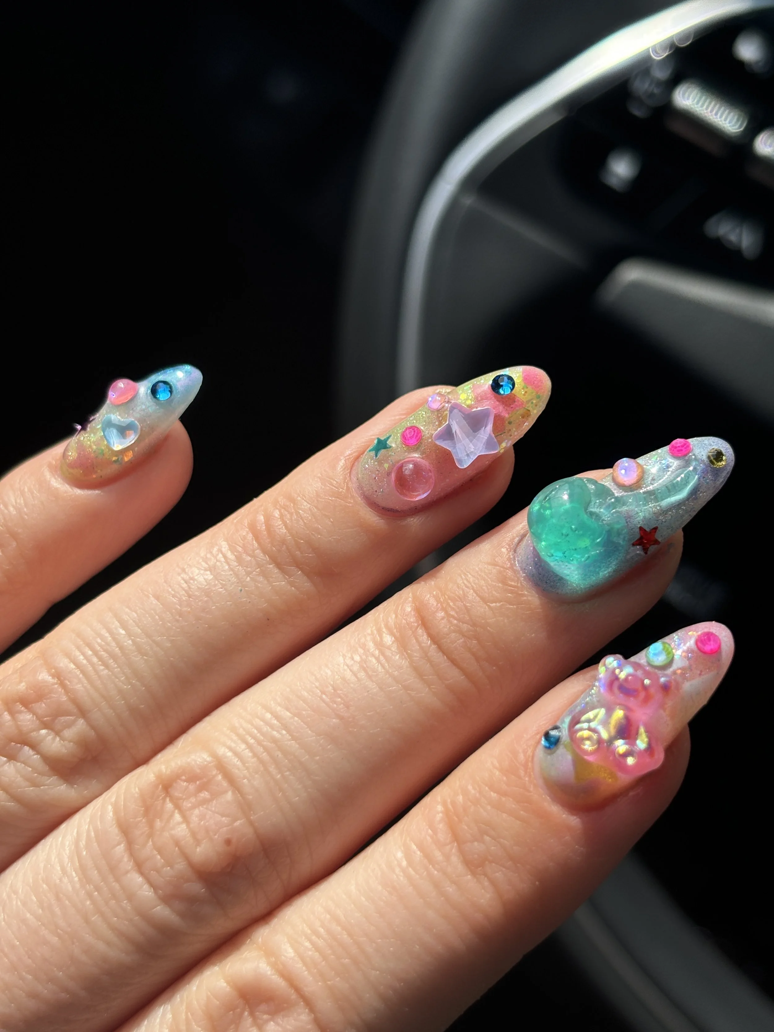 Colorful nails decorated with various small star, heart, and gem embellishments, held over a dark background, with part of a car's dashboard visible in the upper right corner.