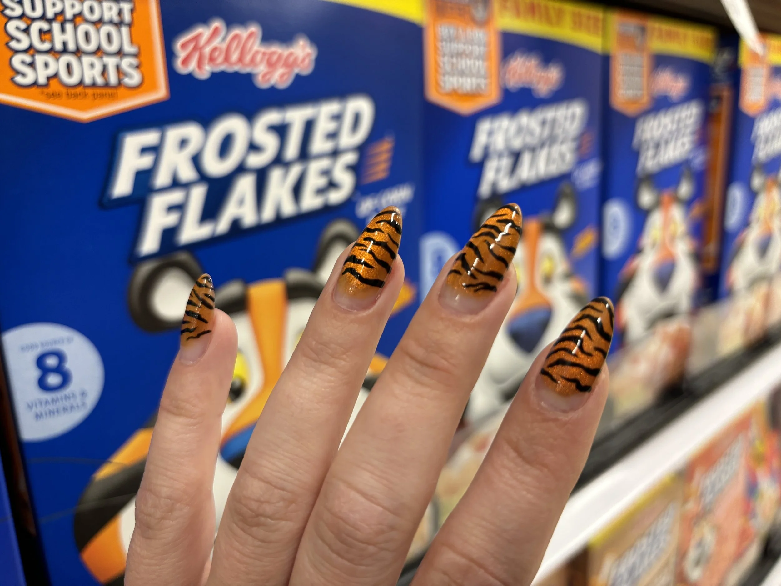 Close-up of a hand with tiger-stripe patterned fake nails in front of a display of Kellogg's Frosted Flakes cereal boxes on store shelves.