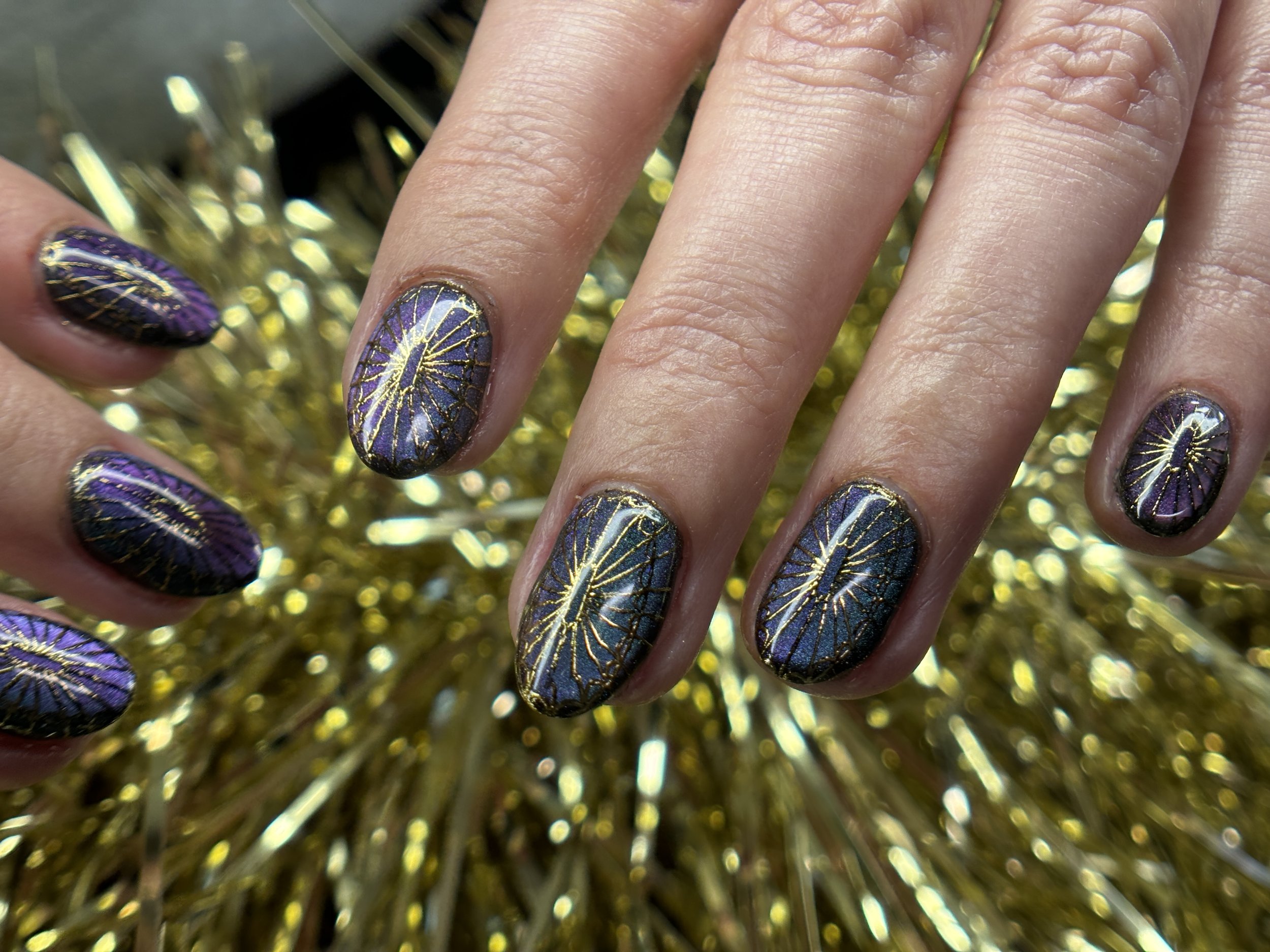 Close-up of decorated fingernails painted with purple and black nail polish featuring intricate gold geometric designs. The background is blurred with gold tinsel.
