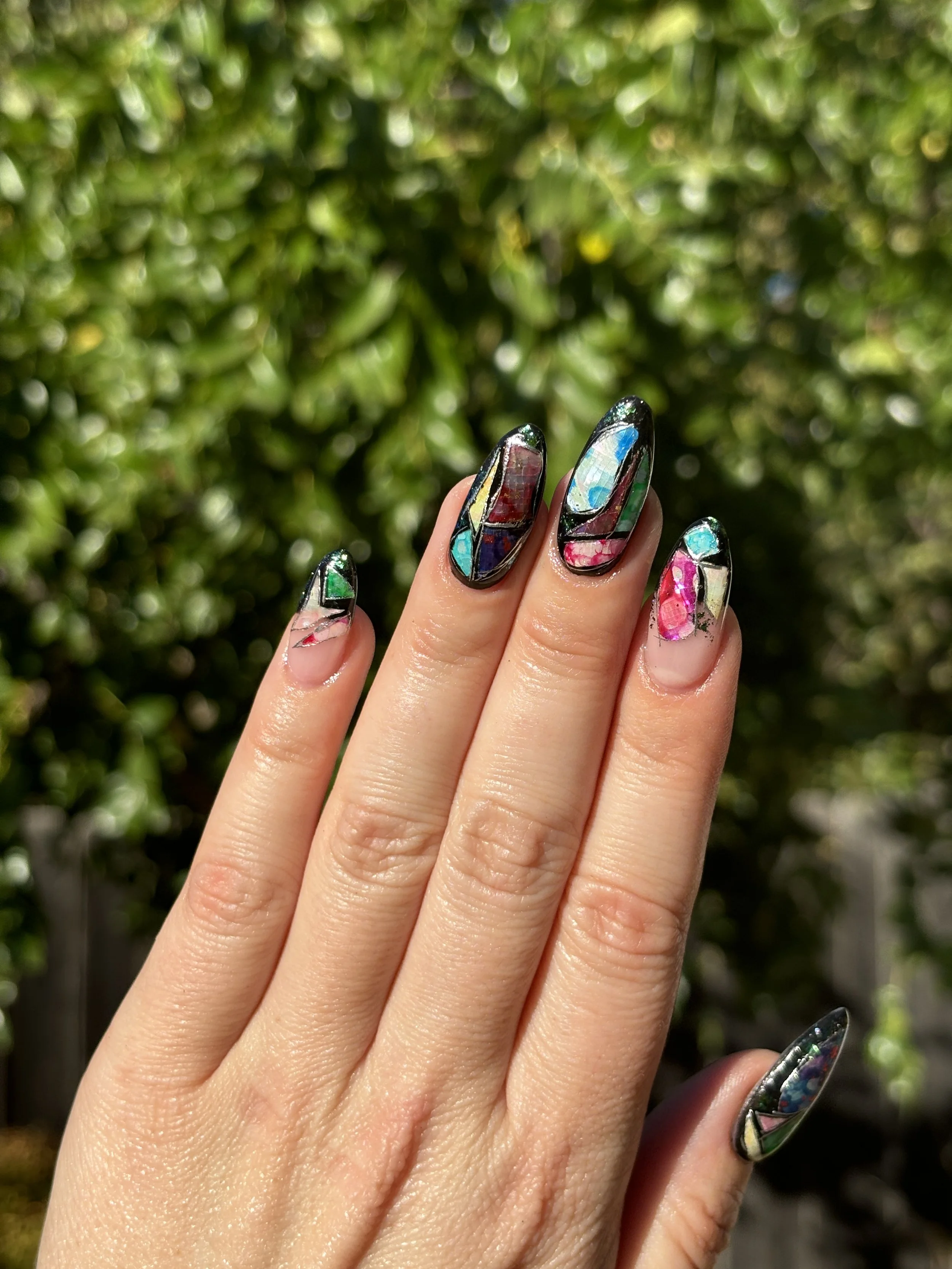 Close-up of a hand with colorful mosaic nail art, set against a blurred green foliage background.