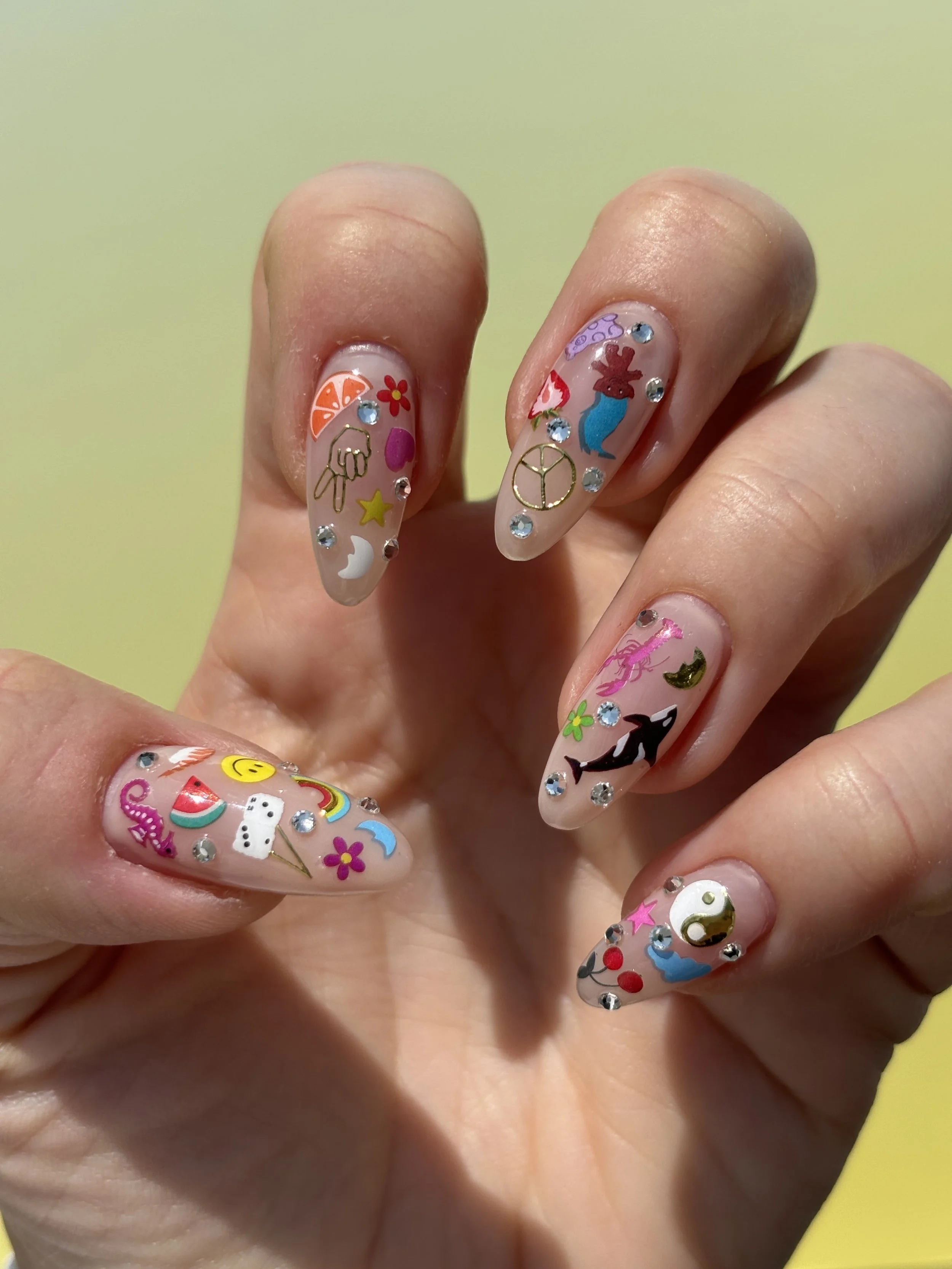 Close-up of a hand with long, decorated nails painted with clear polish featuring stickers, glitters, and rhinestones. The nail art includes colorful images of a unicorn, peace symbols, stars, flowers, a moon, and other whimsical designs.