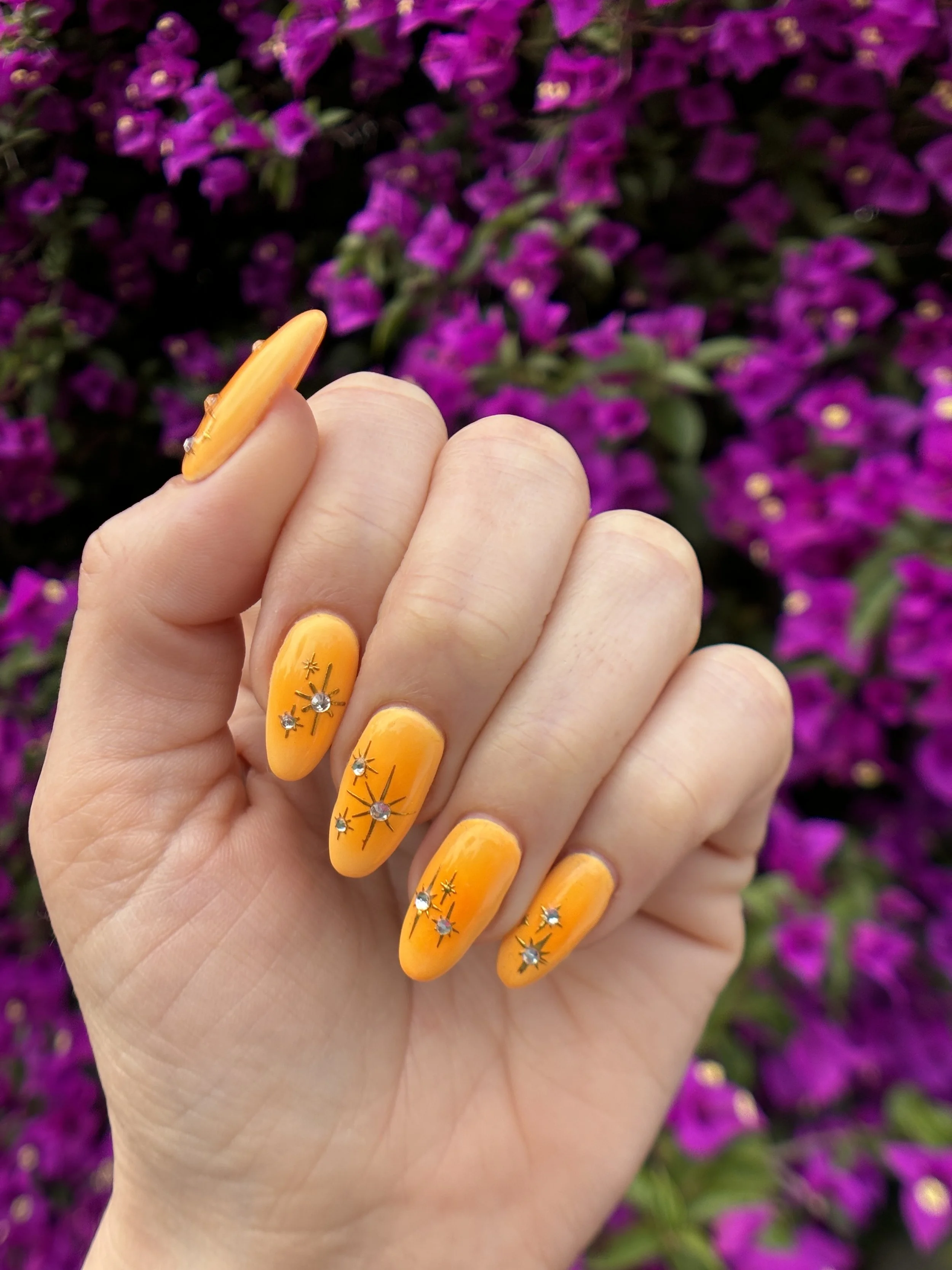 Close-up of a person's hand with bright yellow painted nails adorned with small rhinestone and star-shaped decorations, in front of a background of vibrant purple bougainvillea flowers.