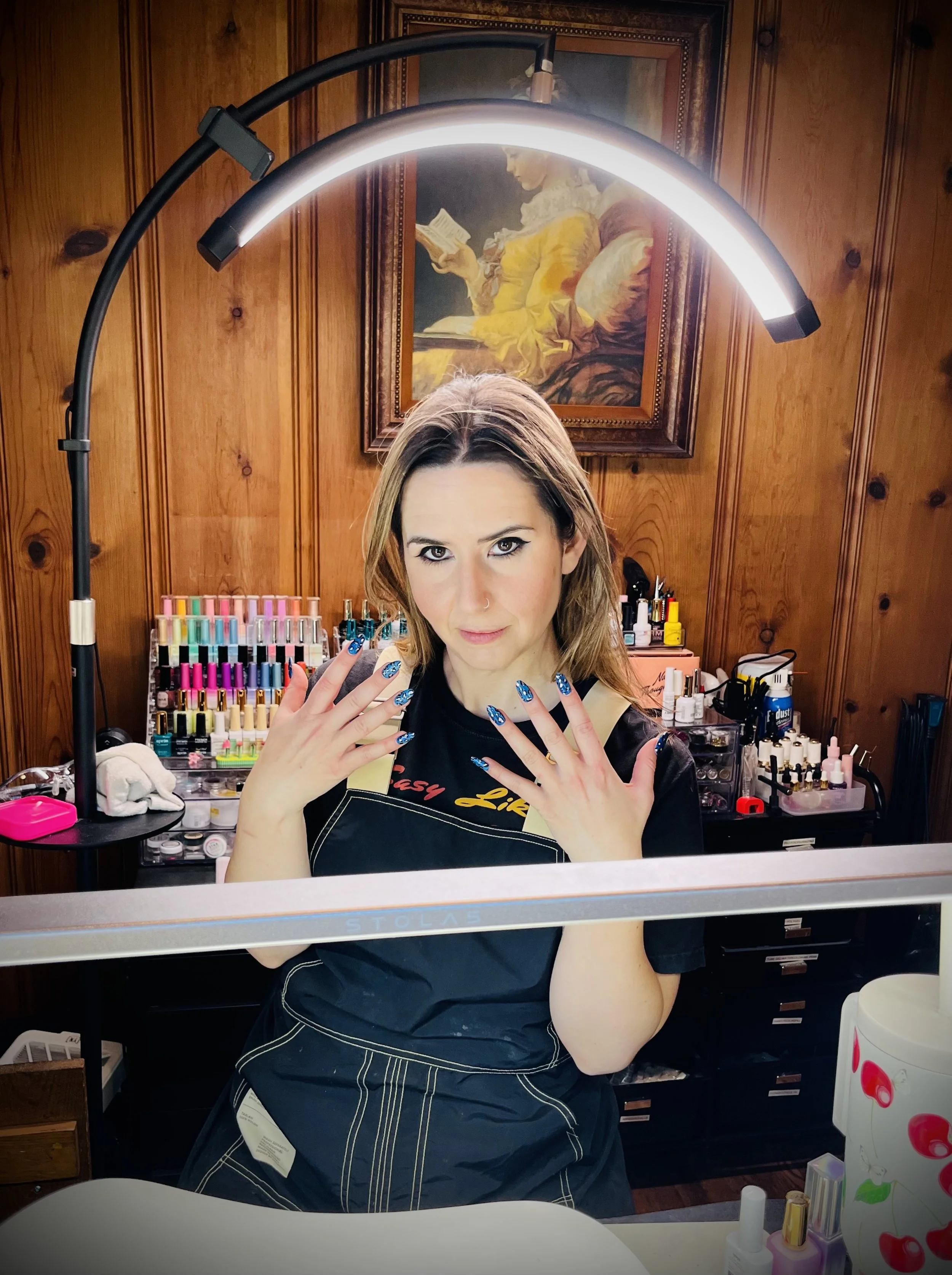 Woman with long brown hair and blue nail polish sitting at a manicure station with bottles of nail polish and supplies behind her. She is looking at the camera with her hands raised, and a mirror with a bright light is overhead. Wooden wall with artwork in the background.