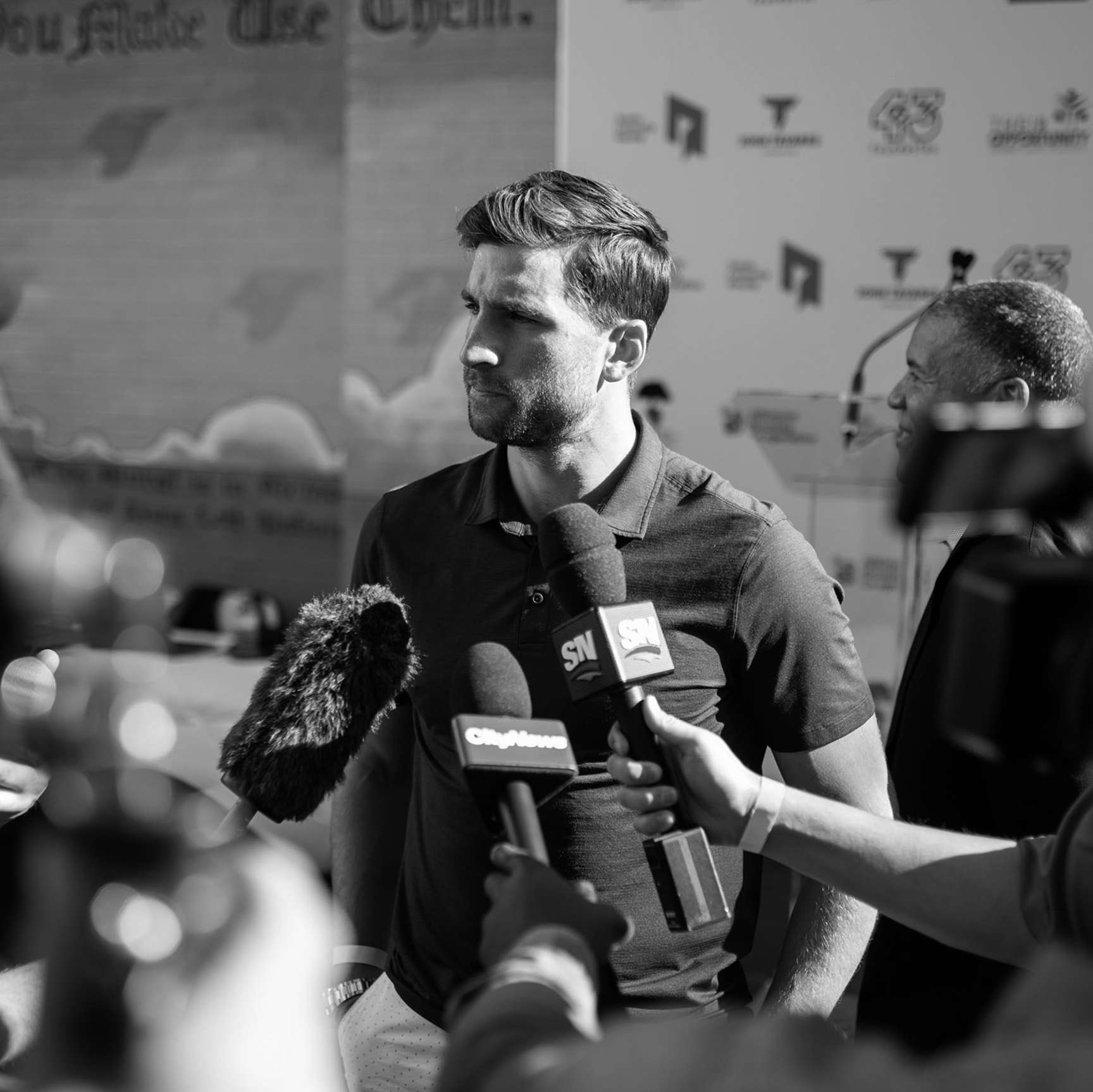 John Tavares being interviewed by multiple reporters and microphones, with a background featuring logos and graphics, in black and white.