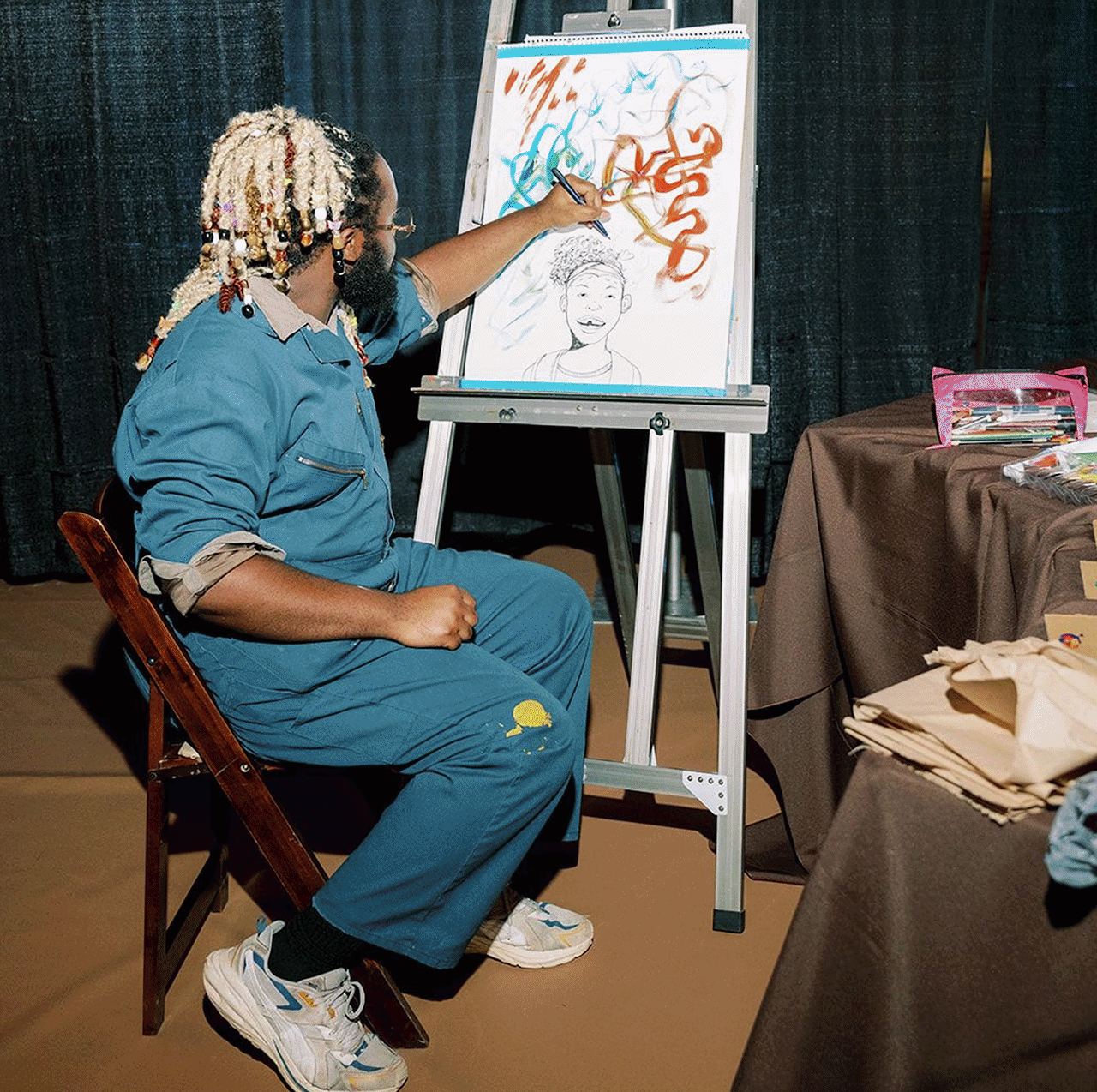 An artist with blonde dreadlocks, wearing a blue jumpsuit, sitting on a wooden chair, painting on an easel. The artwork includes a smiling caricature of a person and colorful abstract designs. On the table nearby are art supplies, including paper and markers, with a bag of supplies on the brown tablecloth.