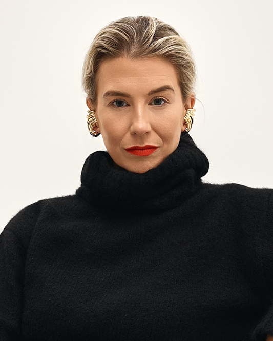 A woman with short blonde hair, wearing a black turtleneck sweater and gold hoop earrings, against a plain white background.