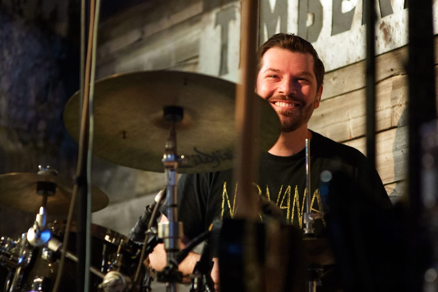 A smiling man with dark hair and a beard playing drums in a wooden room, wearing a black Nirvana T-shirt.