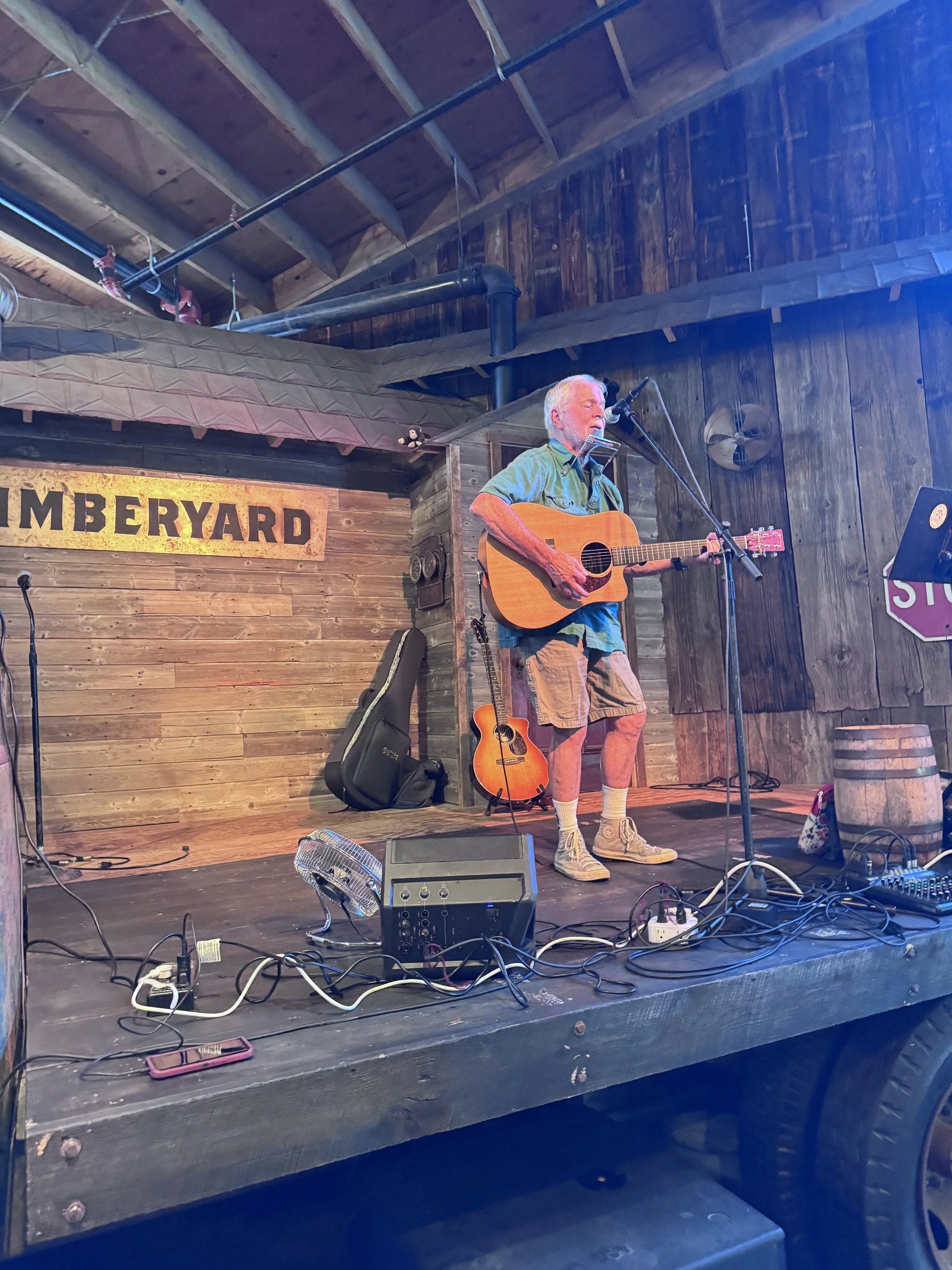 An older man with white hair wearing a green shirt and khaki shorts playing an acoustic guitar and singing into a microphone on a wooden stage at a venue called 'GARDENERYARD.' There are various musical equipment and cables scattered on the stage.