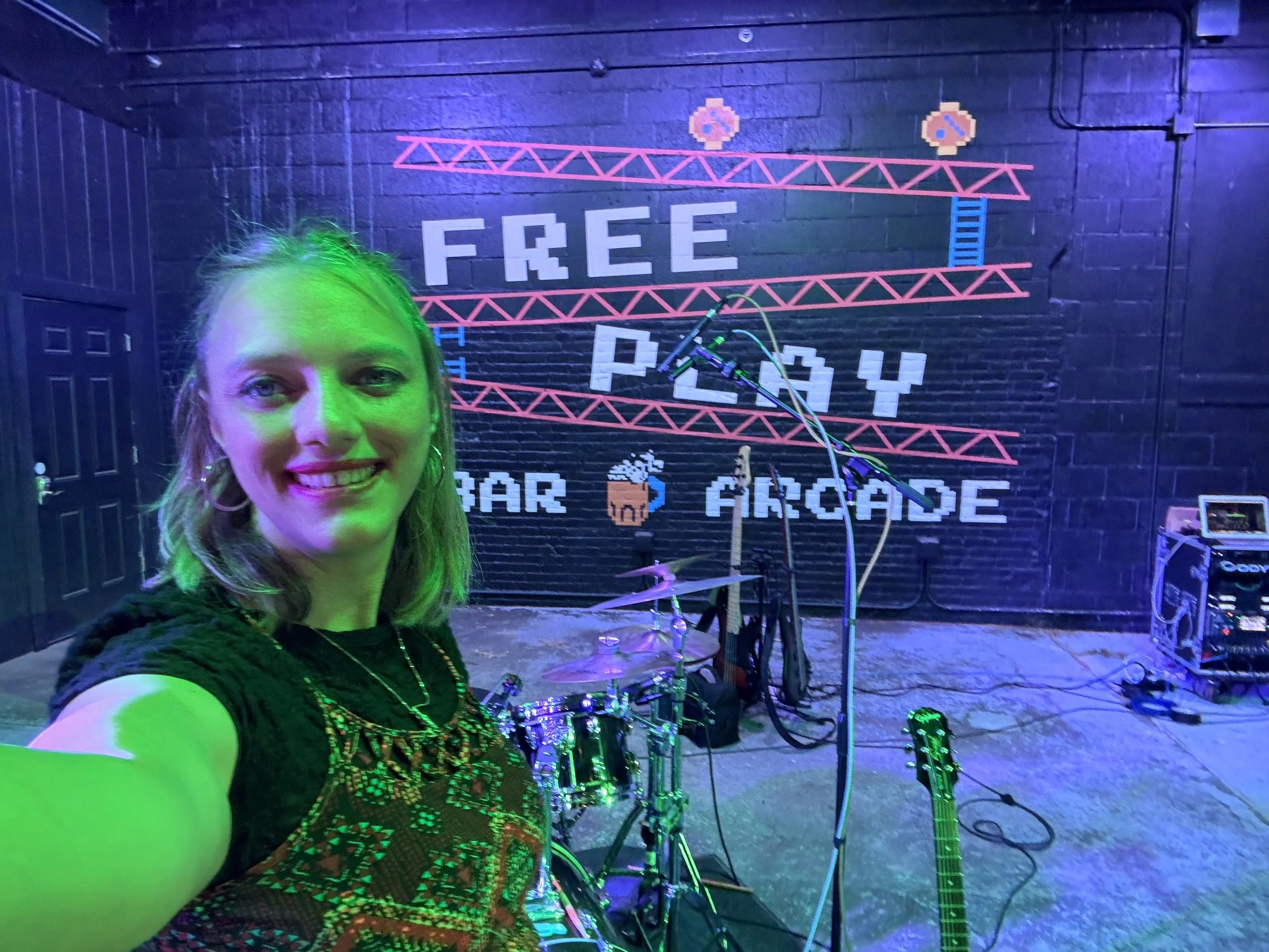 A woman with green hair and a patterned top taking a selfie in front of a mural that reads 'FREE' and 'PAY' on a black brick wall, with pixel art style graphics and electronic equipment.