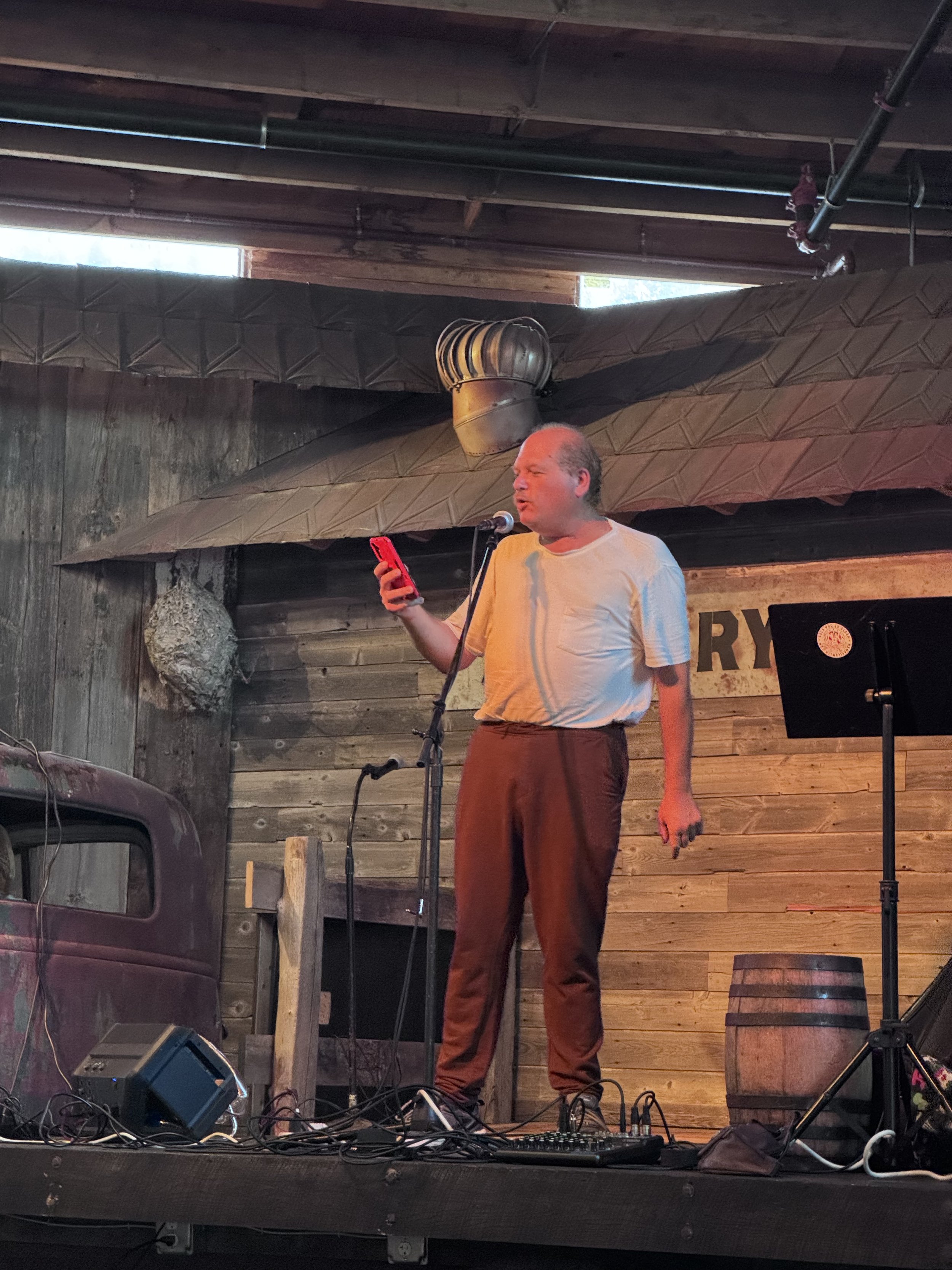 Man singing into a microphone while holding a red phone, standing on a stage with wooden wall background and musical equipment, including a piano and a barrel.