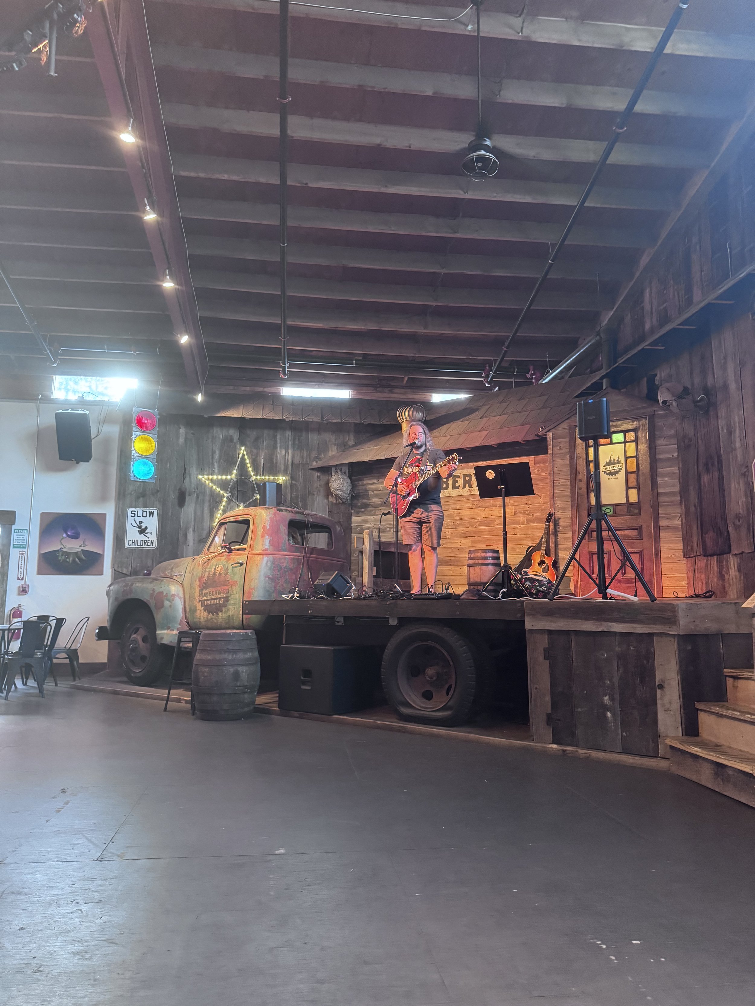 A man playing guitar on a small stage made from a flatbed truck with a rusted vintage car on the front, in a rustic indoor setting with wooden walls, stage lights, and a hanging star-shaped light decoration.