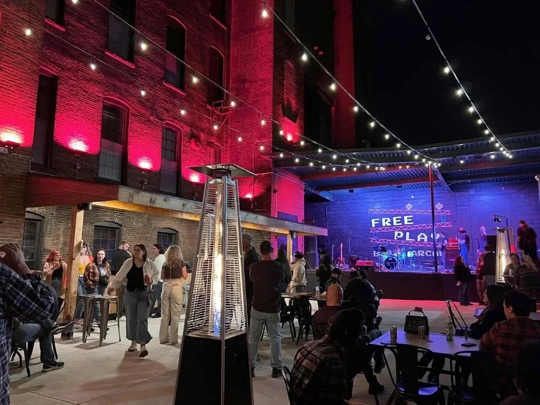 Outdoor concert venue at night with a stage, string lights overhead, and people socializing. The stage has a sign that reads 'FREE PLAY' and 'BRANDI MERCI'.