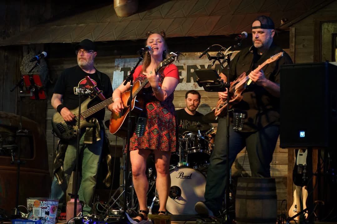 A four-member band performing on stage with musical instruments in a rustic wooden venue.