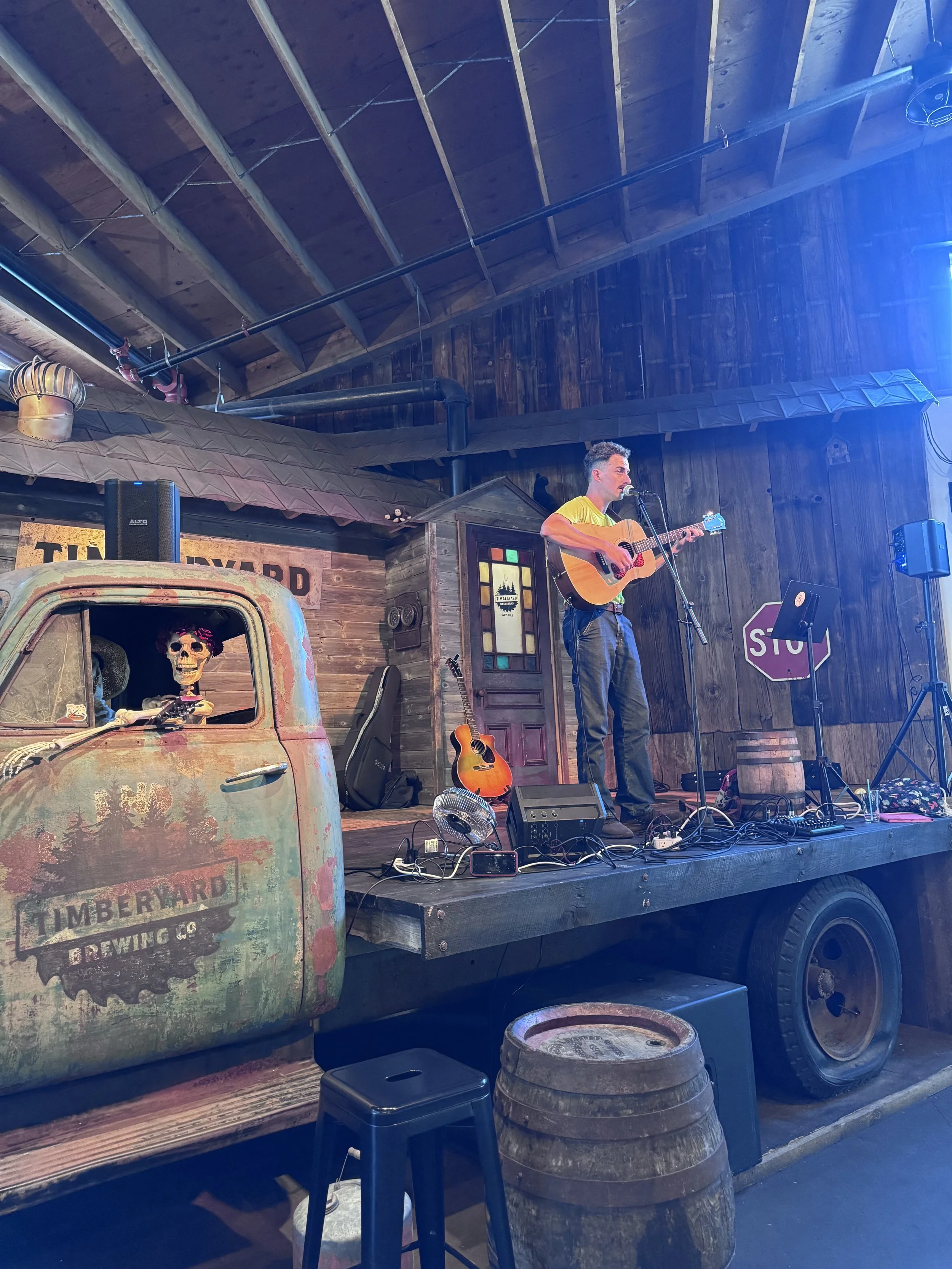 A musician playing an acoustic guitar and singing into a microphone on a stage inside a rustic venue with wooden walls and a high ceiling. There are musical equipment, a guitar, and decorative items including a skeleton figure in a vintage vehicle.