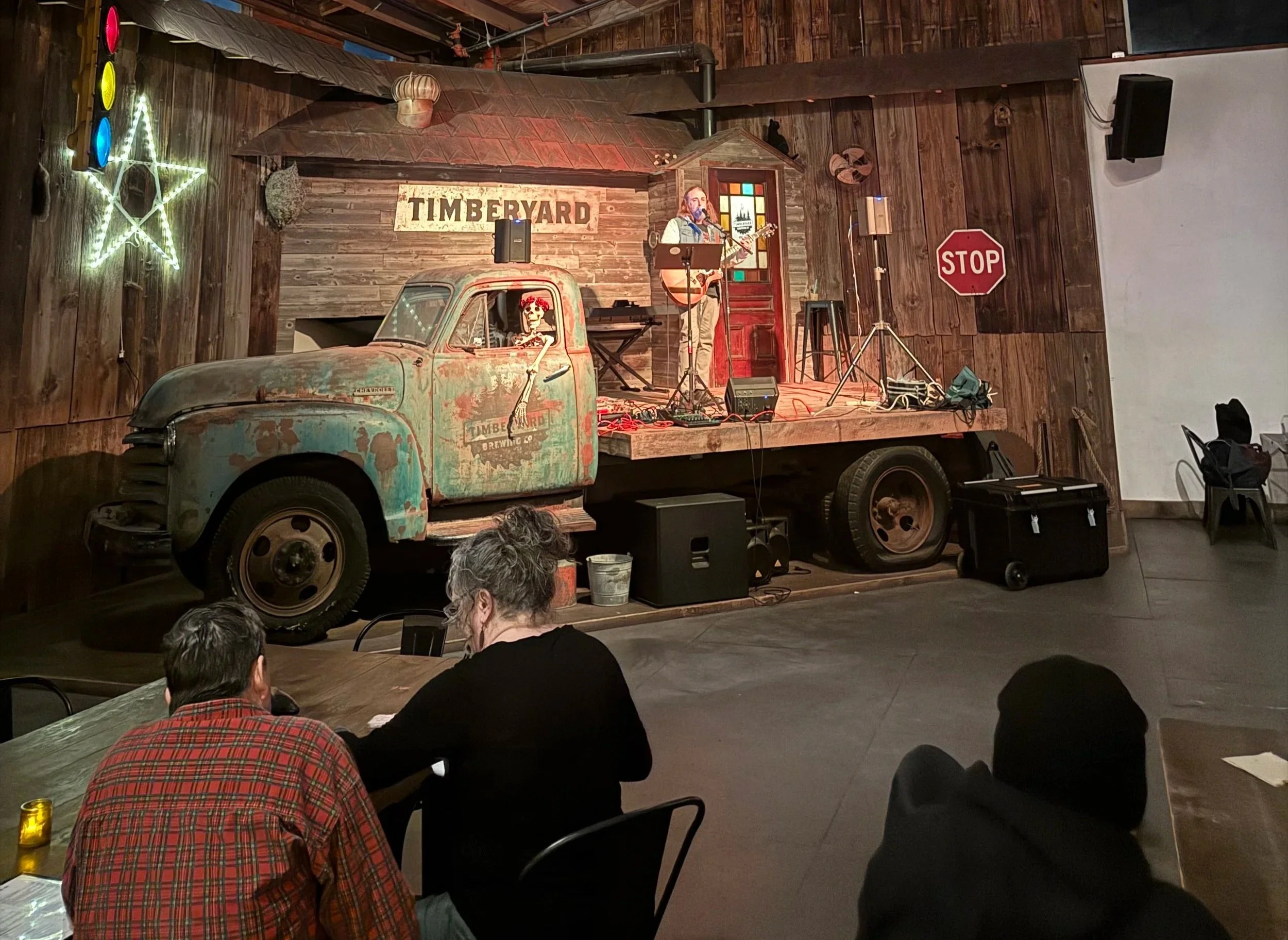 Indoor venue with a flatbed truck stage decorated rustic, featuring an illuminated star and traffic light, a skeleton figure in the truck cabin, and a musician performing with a guitar, surrounded by posters, sound equipment, and audience seated at tables.