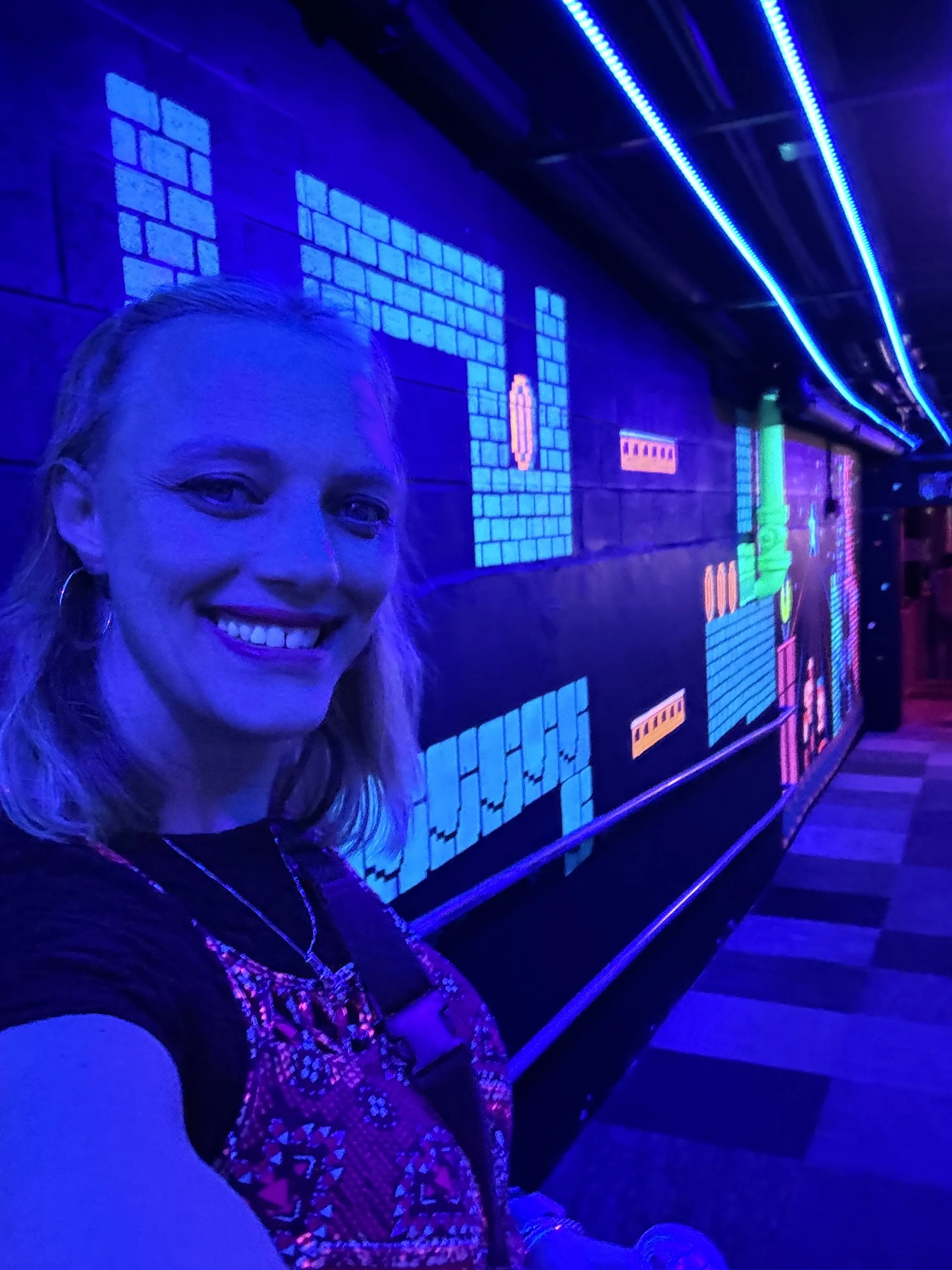 A woman with blonde hair and glasses taking a selfie in a dark room with vibrant blue and purple neon lights and digital pixel designs on the wall behind her.