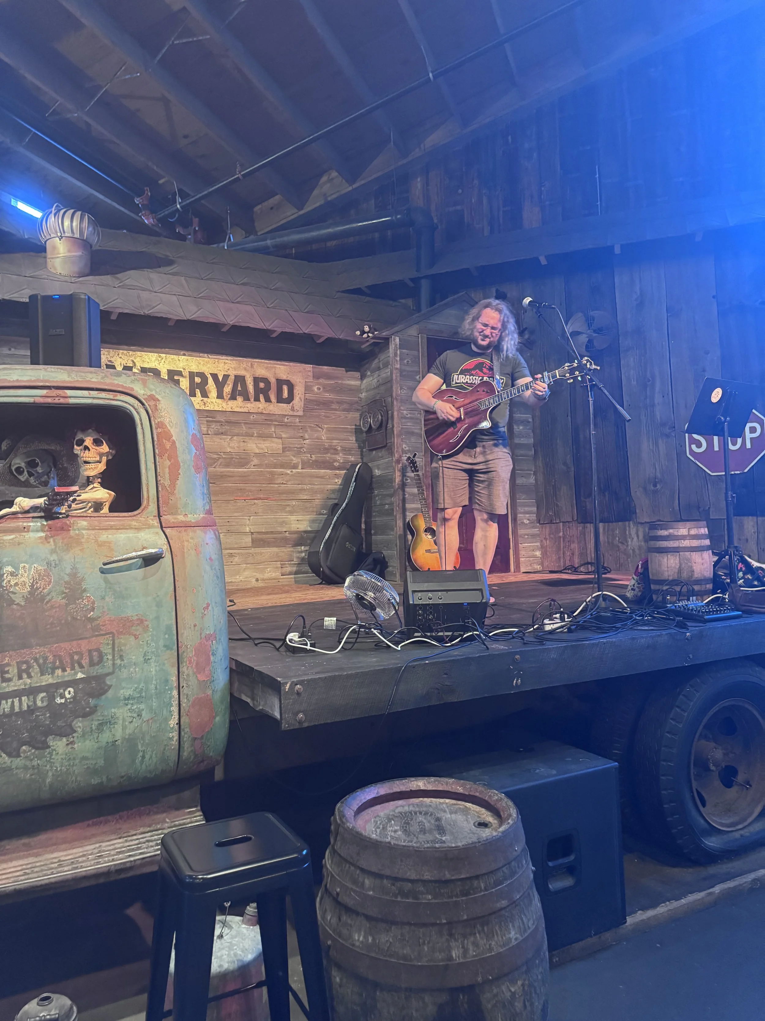 A person playing guitar on a small stage with a wooden backdrop that has a 'Greeneryard' sign. There is a vintage truck with skull decorations and a toy horse on top, and various stage equipment including a microphone, music stand, and speakers.