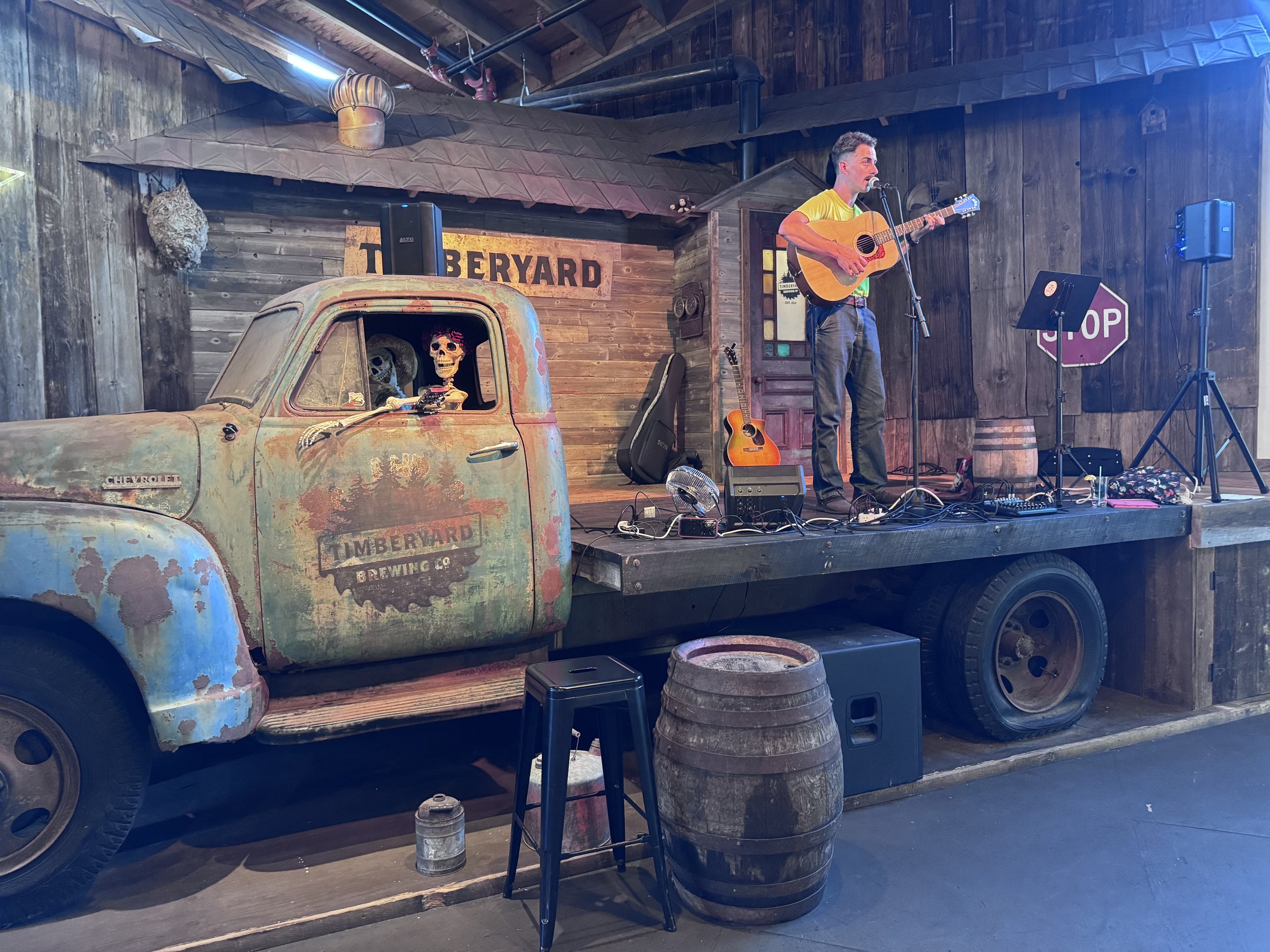 A rustic stage setup inside a wooden building with a man playing guitar and singing into a microphone. The stage has a vintage Chevrolet truck with skeleton decorations in the cab, a sign reading 'Timberyard,' a couple of guitars, and various equipme