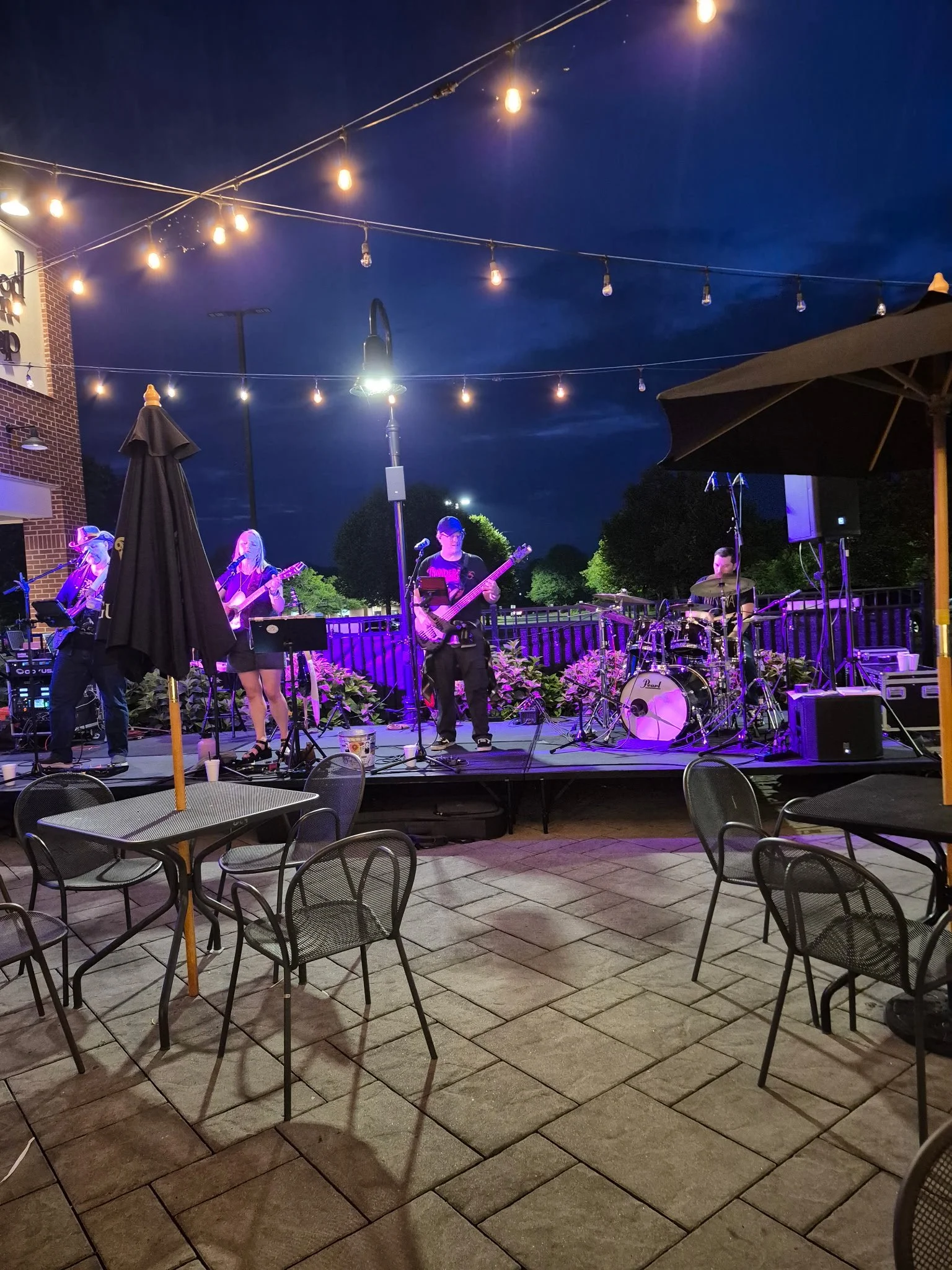 An outdoor stage with a band performing under string lights at night. There are tables and chairs in the foreground.