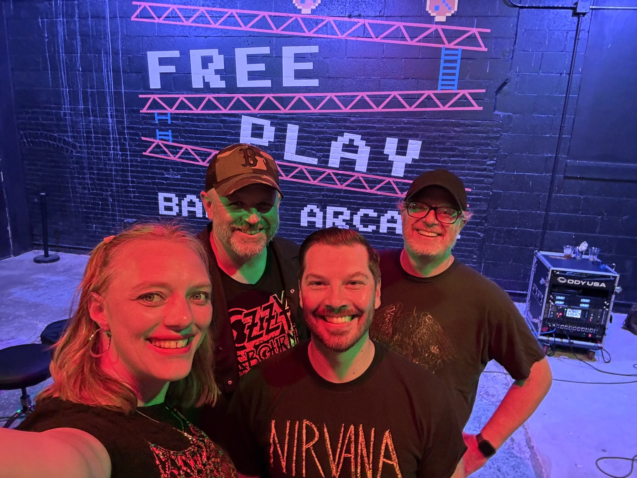 A group of five people smiling at a party or event, with a black brick wall behind them displaying neon-colored pixelated text that says 'FREE PLAY' and other smaller words, with some event equipment visible to the side.