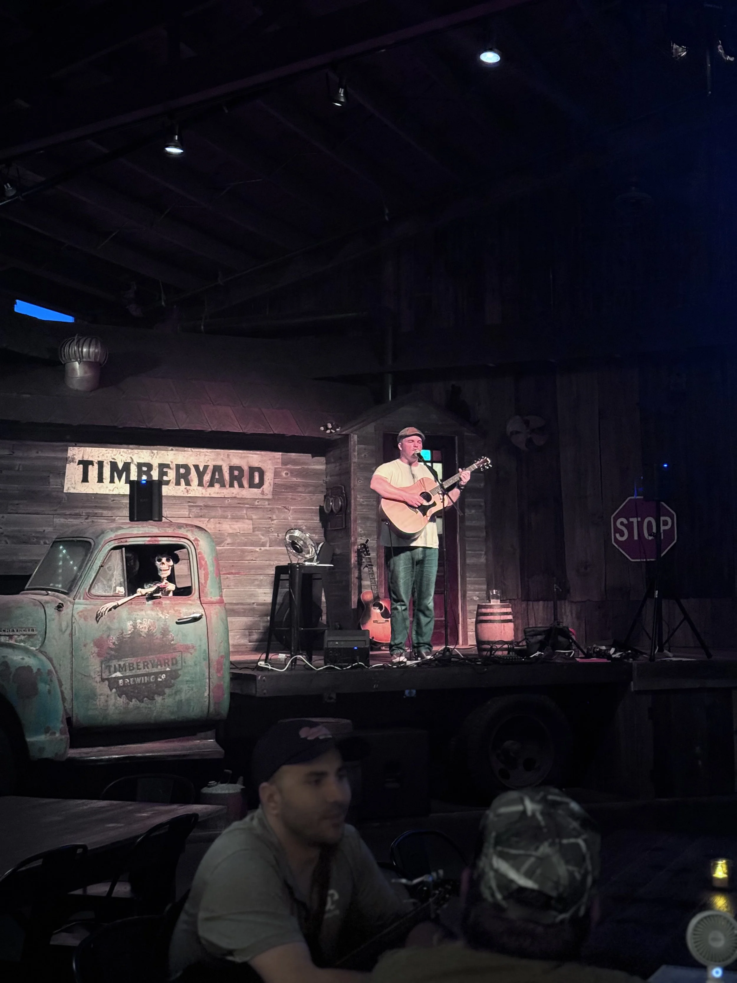 A man performs a song with an acoustic guitar on a stage at Timberyard Brewing Co., with a small audience watching.