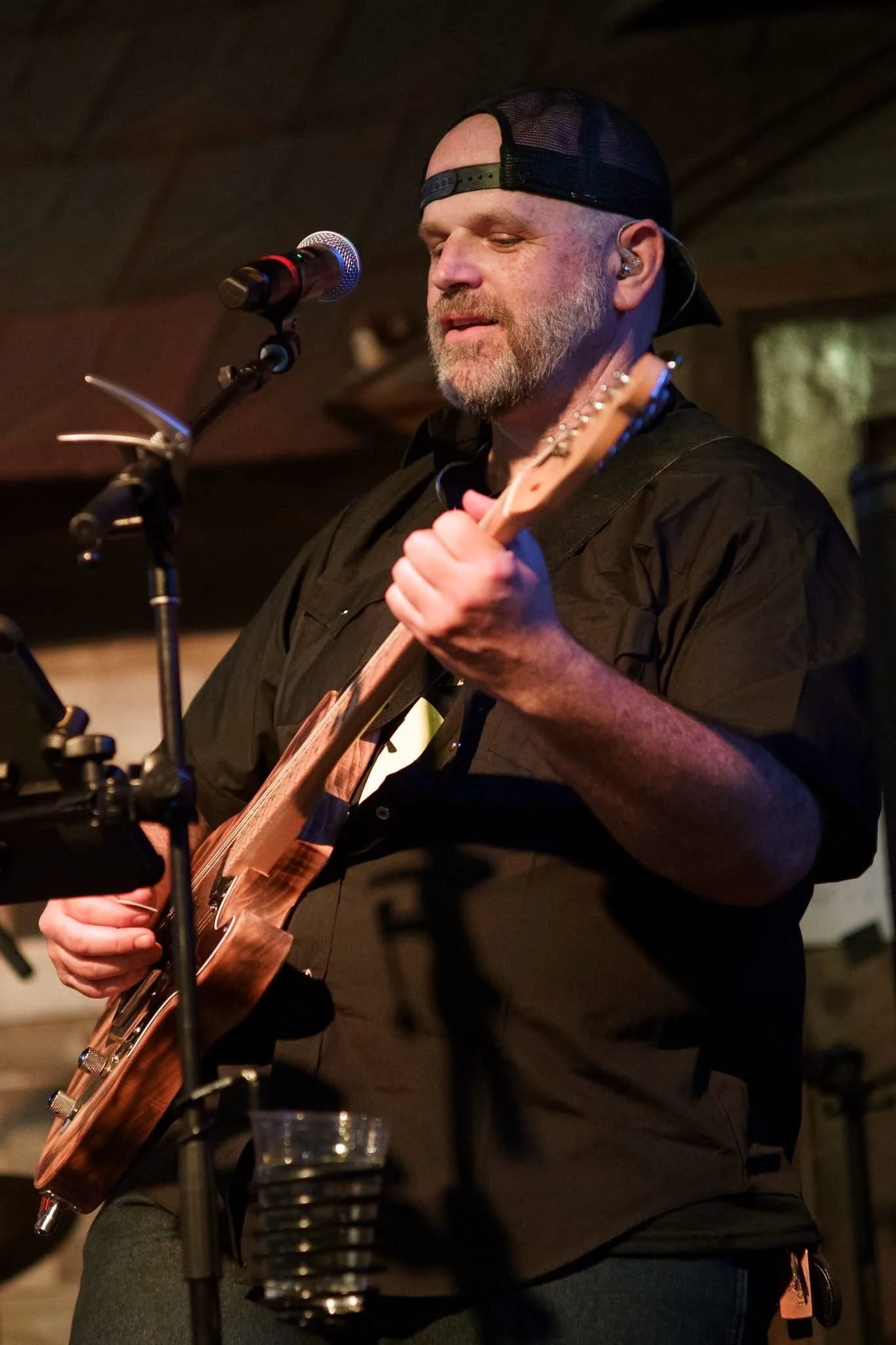 A man with a beard wearing a black cap backwards, playing an acoustic guitar and singing into a microphone in a dimly lit indoor setting.