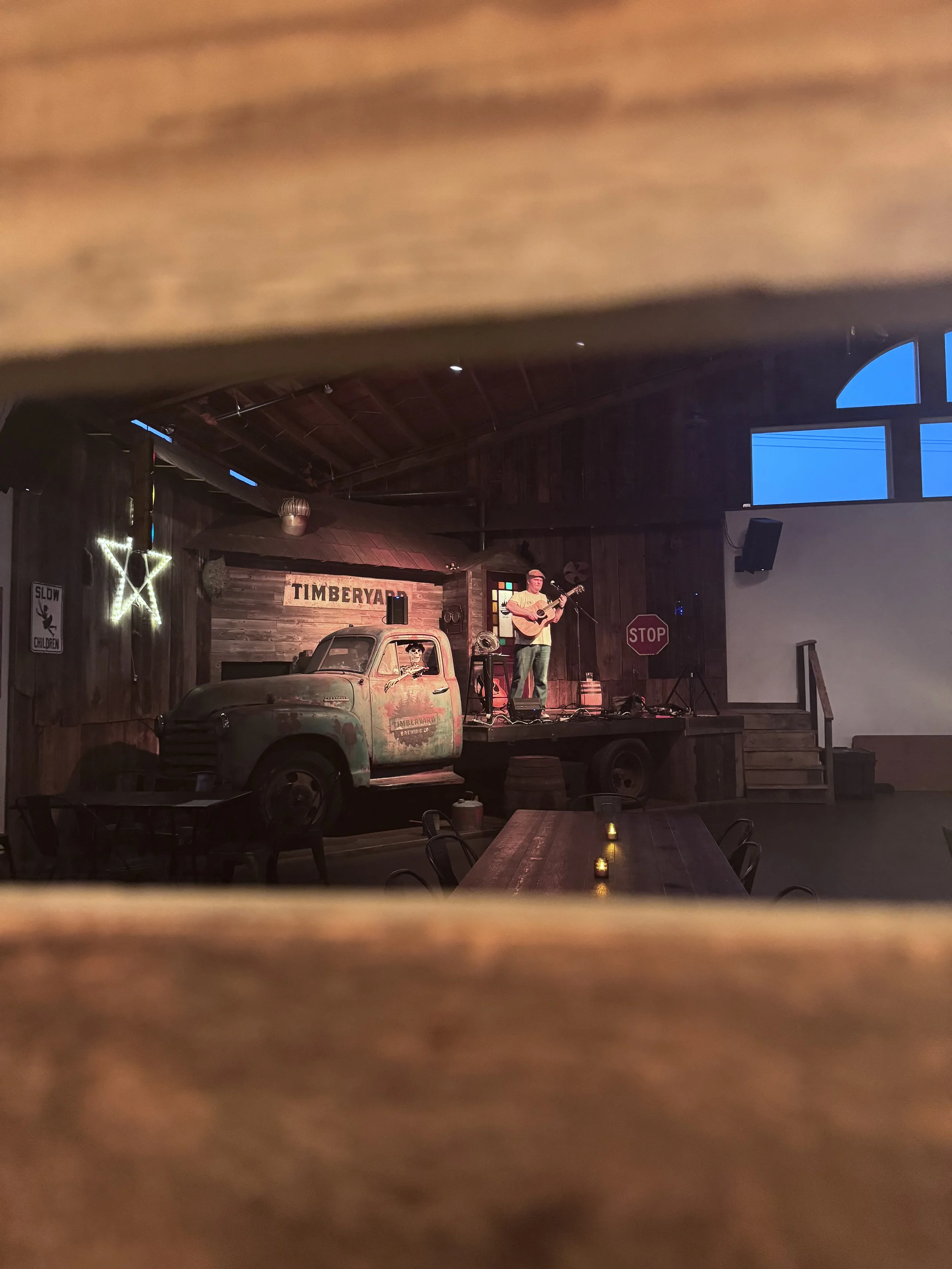 A musician playing a guitar on a small stage with a vintage truck and wooden sign that says 'Timberyard Brewing Co.' in the background, inside a rustic venue with wooden walls and decor.