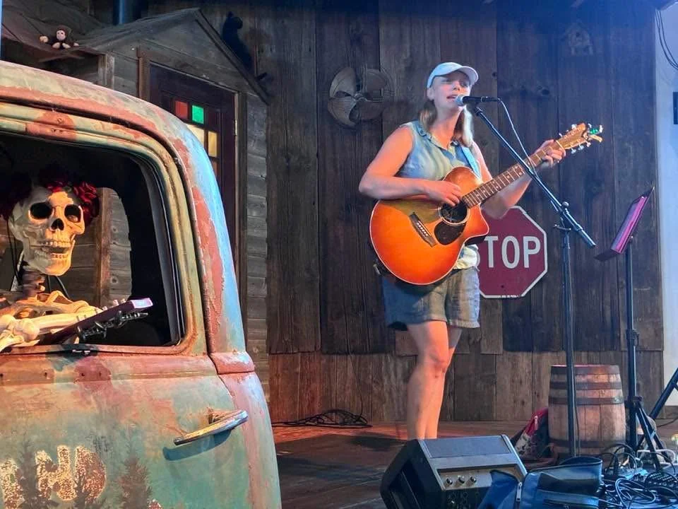 A woman with a guitar singing into a microphone on stage, in a rustic setting with wood paneling, a STOP sign, and a skeleton in a rusted truck nearby.