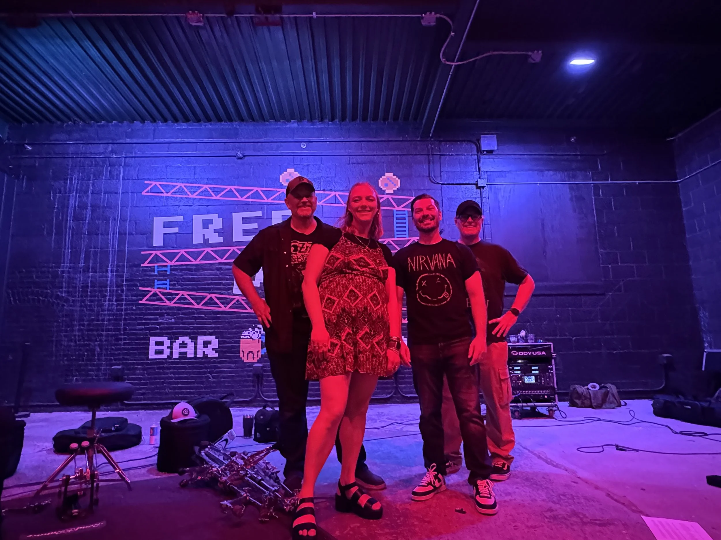 Four people standing together on a stage in front of a neon sign, with DJ equipment and musical gear on the floor, and a dark blue brick wall behind them. The lighting is colored with purple and pink hues.