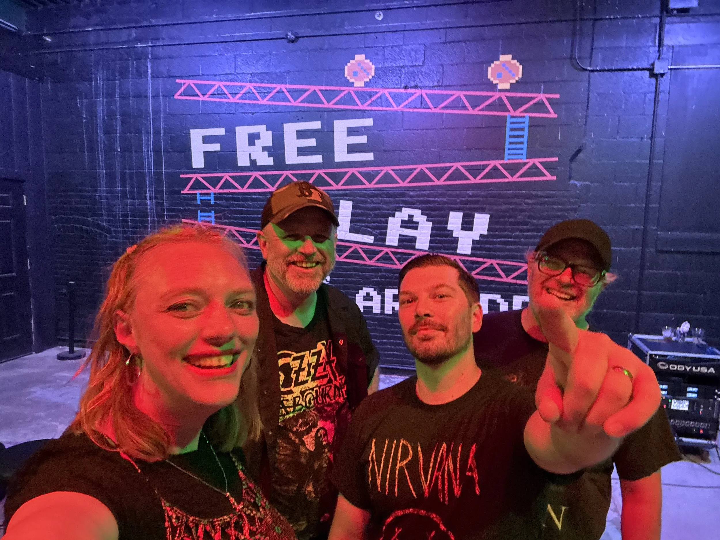 Group of five people taking a selfie at an indoor event with a blue brick wall in the background. The wall has pixel art of two hearts and the word 'FREE' in large letters. The people are smiling and appear to be enjoying themselves.