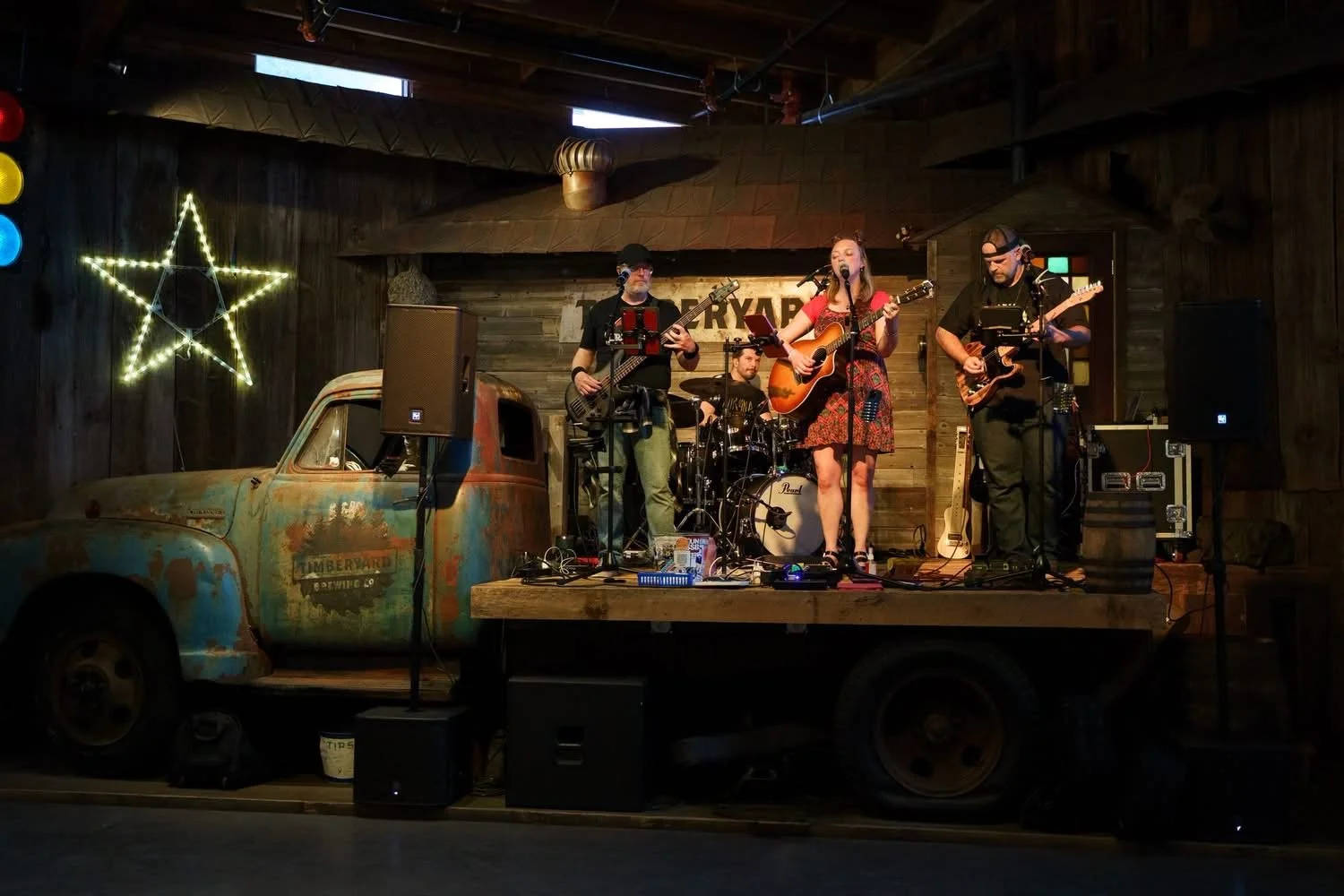 A four-piece band performing on a rustic stage with a vintage truck, a star-shaped light, and wooden walls. The band includes a woman playing guitar and singing, and three men playing guitar and drums.