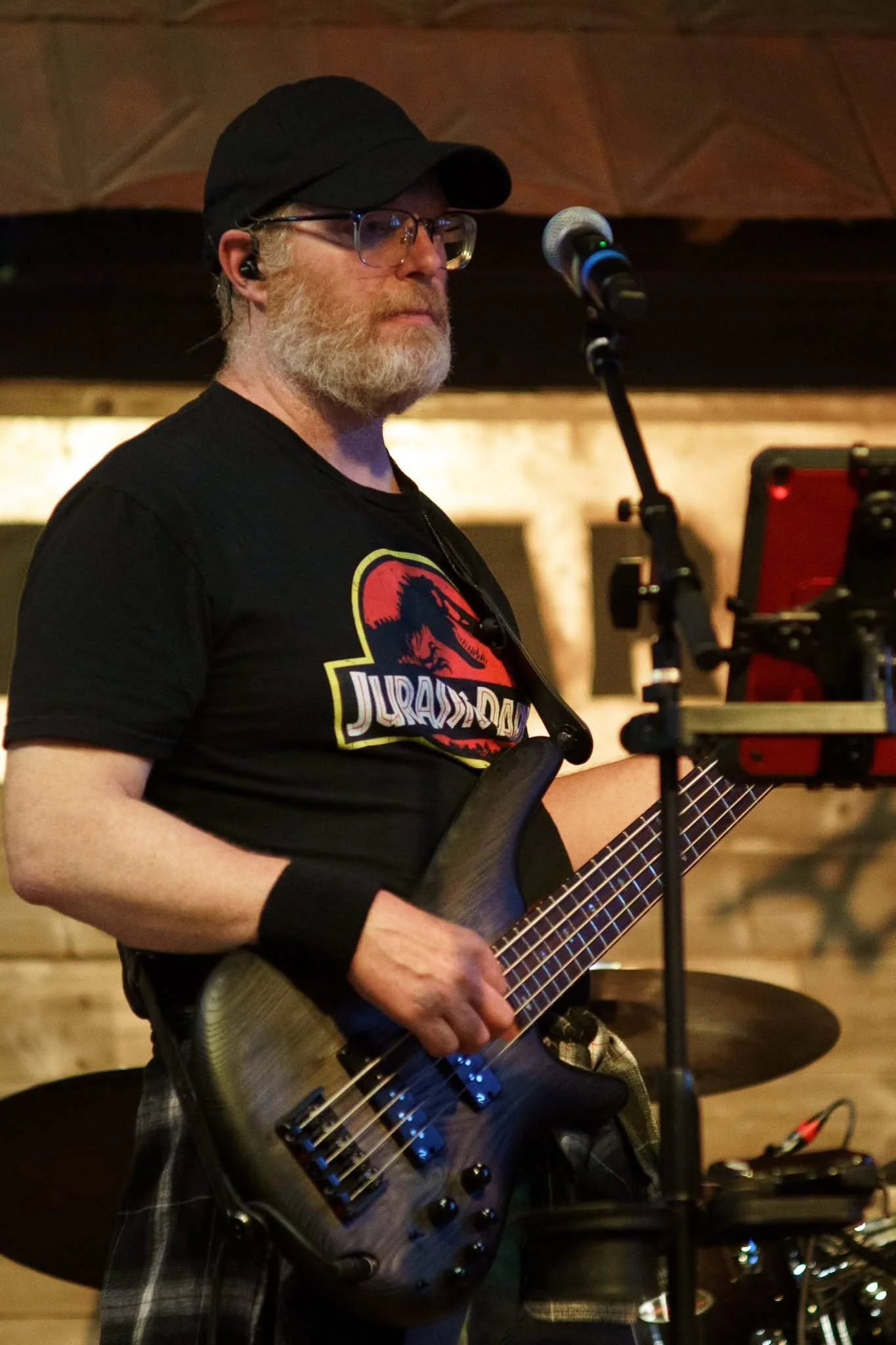 A man with glasses, a black cap, and a beard playing an electric guitar at a music venue, with a microphone and drum set nearby.