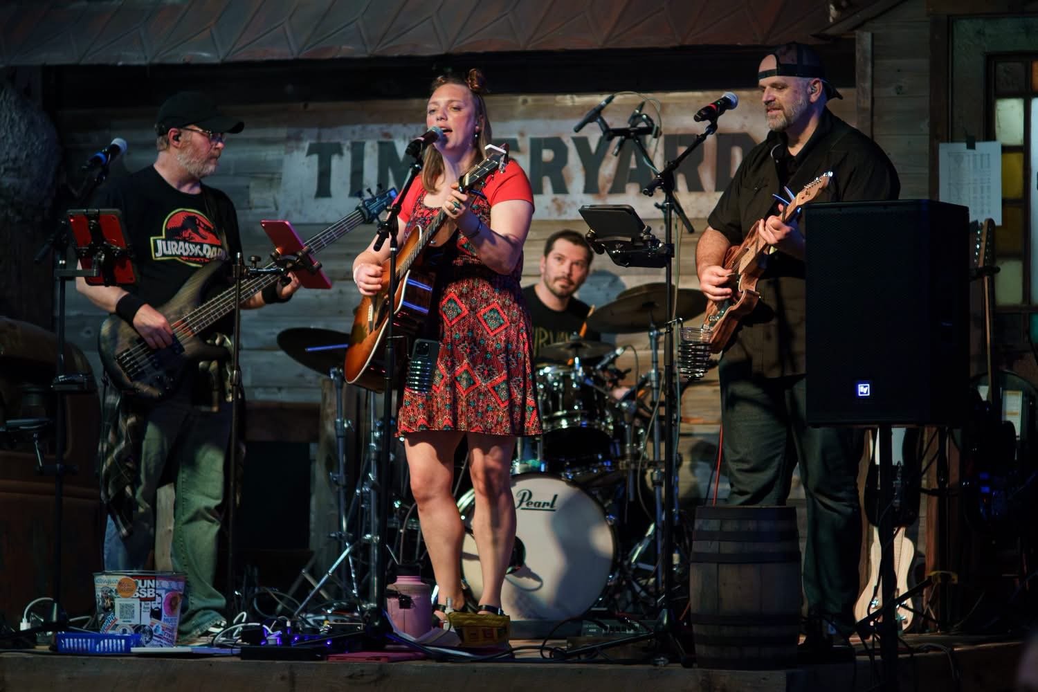 A band performing live on stage at a venue called Timberyard. The band includes a woman singing and playing guitar, two men on guitar and bass, and a drummer in the background.