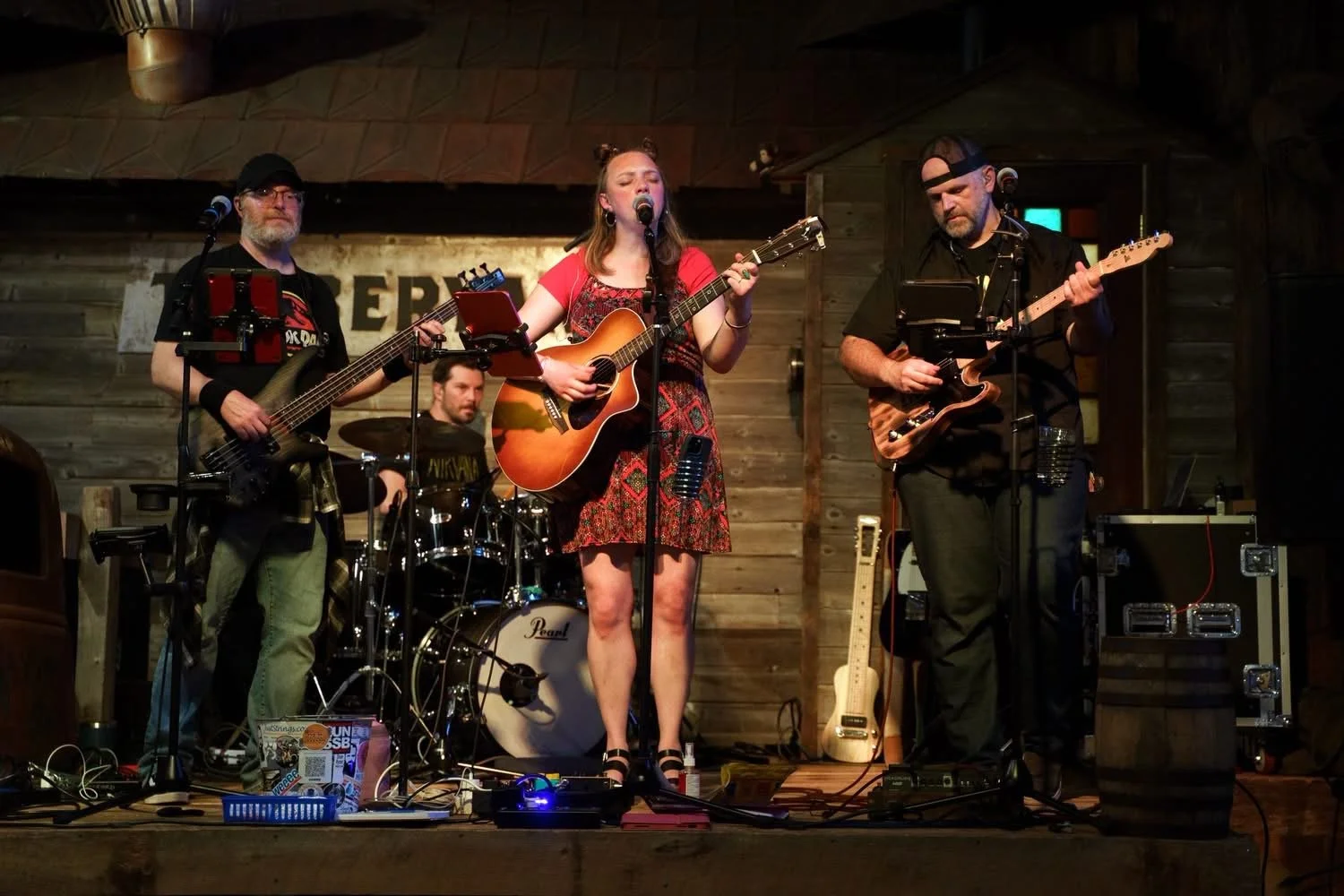 A four-member band performing on a wooden stage in a rustic venue. The woman in the center is singing and playing an acoustic guitar, flanked by two men playing electric guitars, and a drummer in the background.