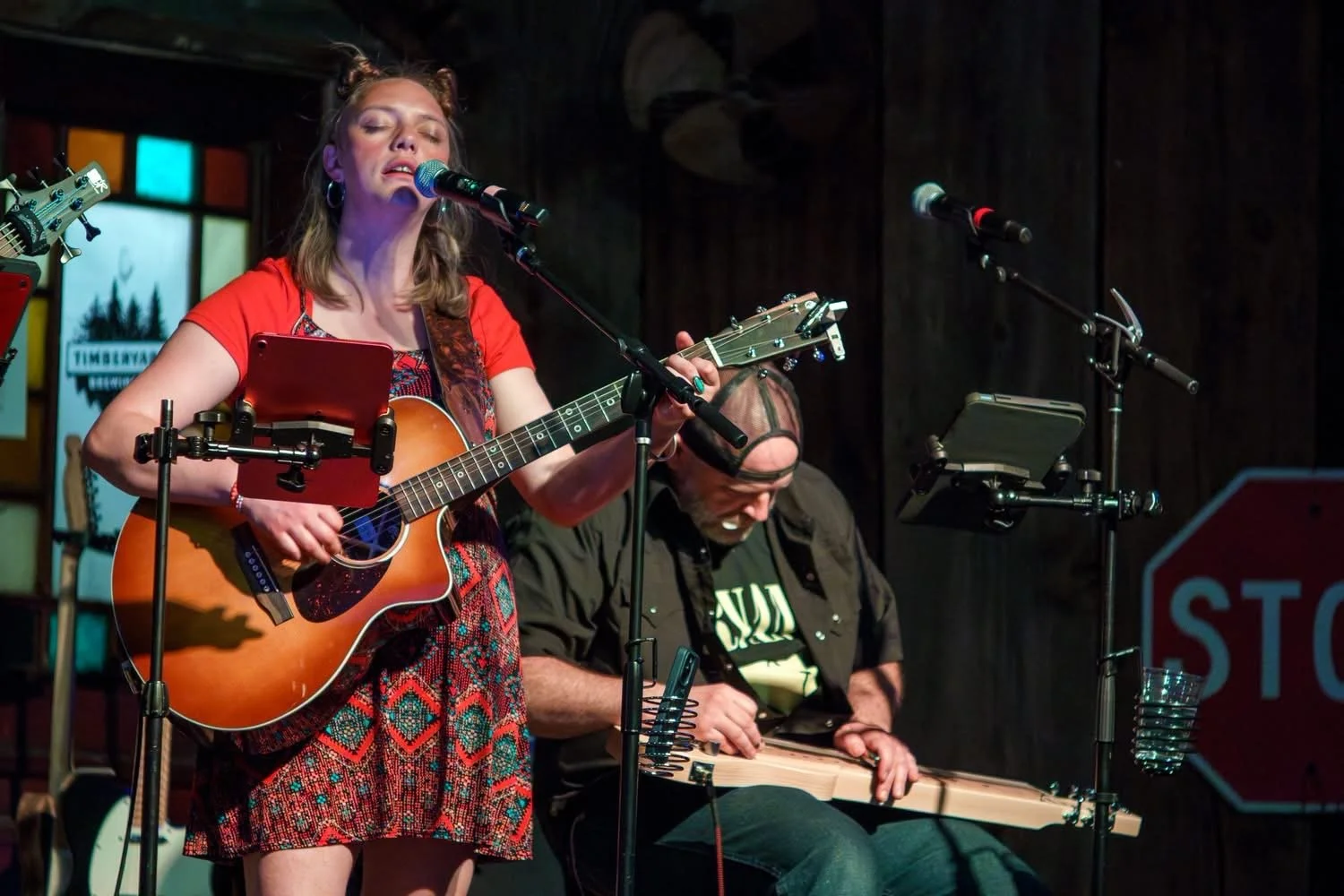 A woman singing and playing an acoustic guitar on stage with a man playing a lap steel guitar behind her.