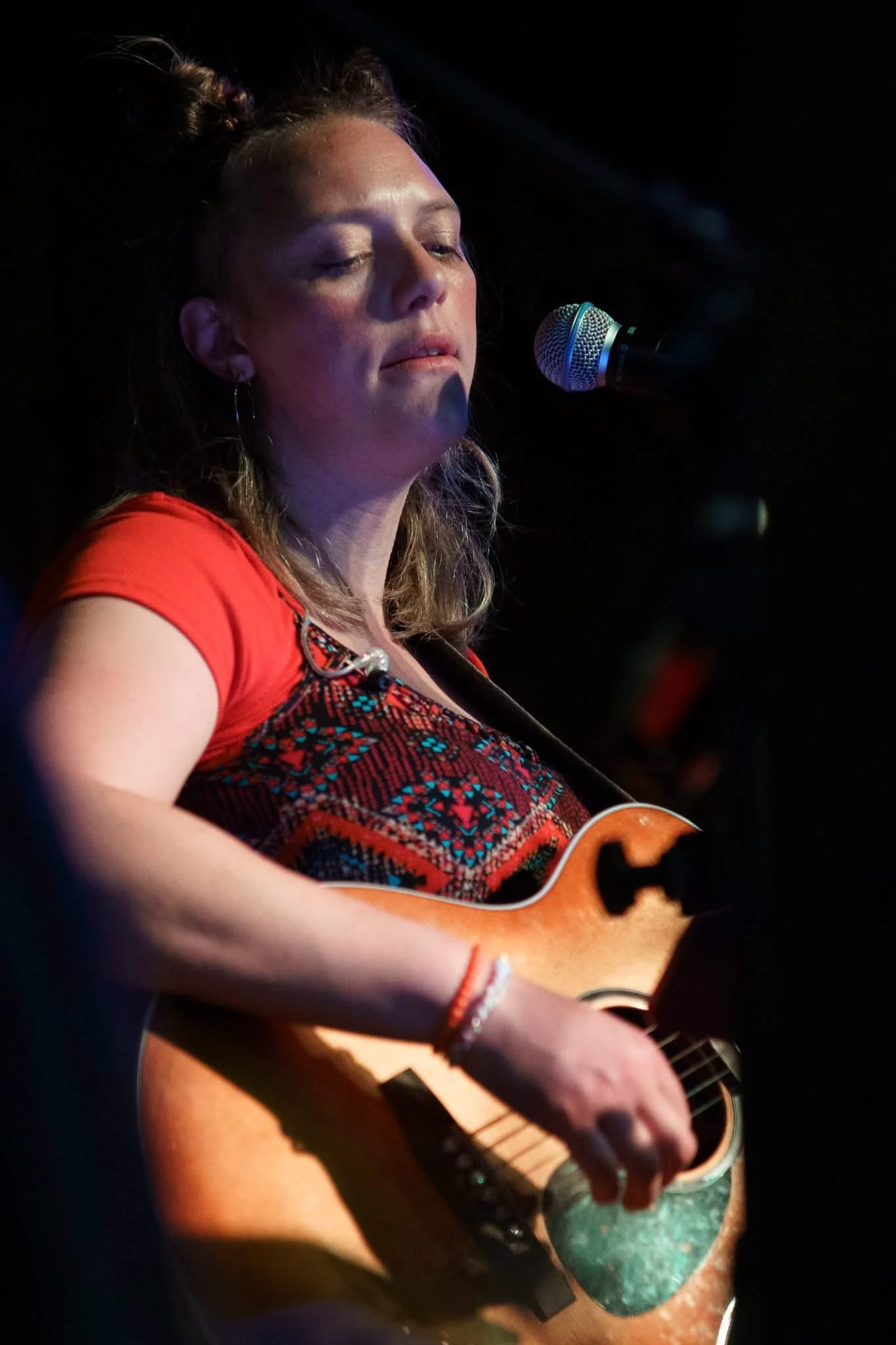 A woman singing into a microphone while playing an acoustic guitar on a dark stage.