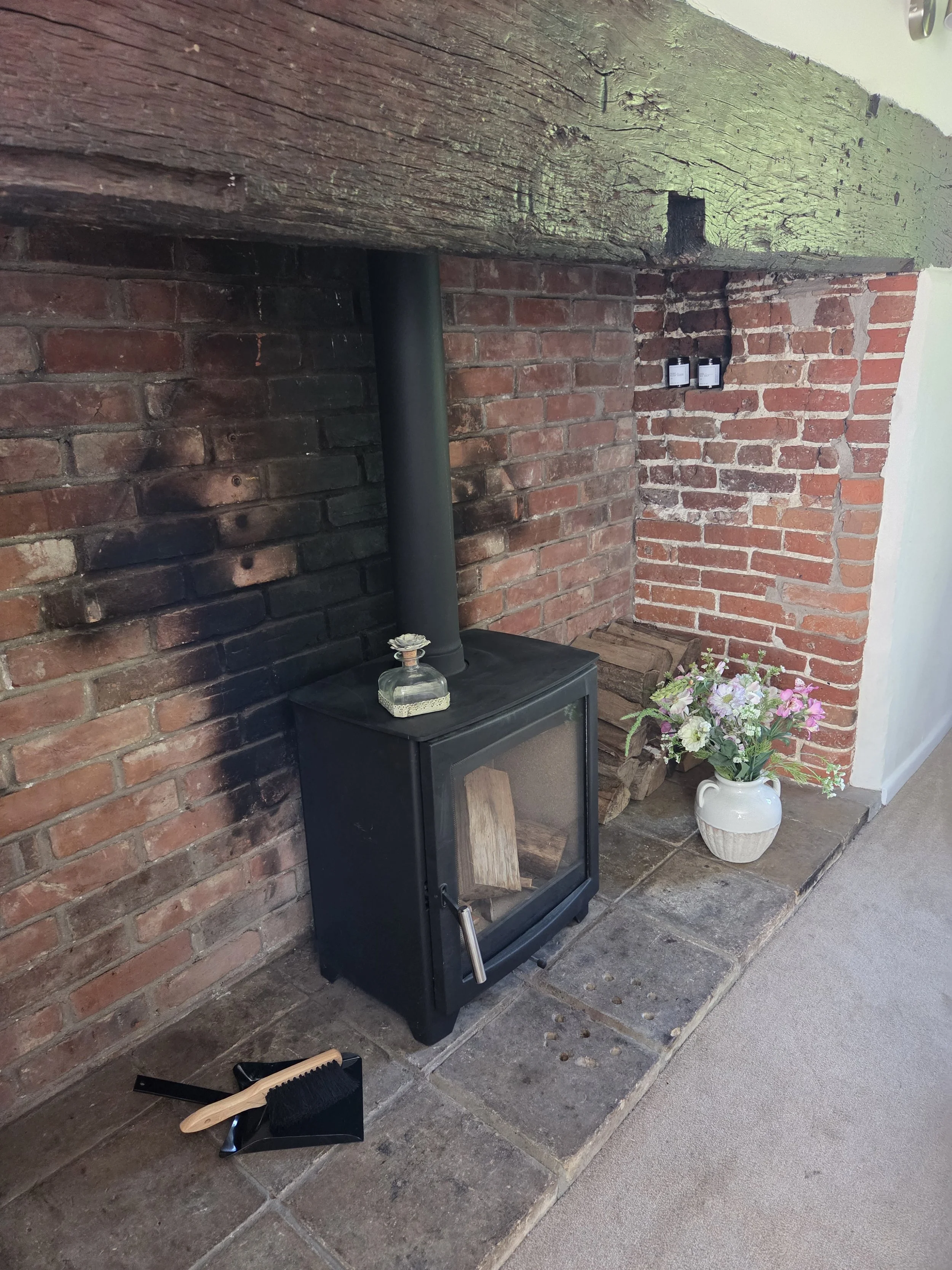 A black wood-burning stove with a glass door and stacked firewood inside, situated against an exposed brick wall, with a brass decorative item on top, a bouquet of pink and white flowers in a white ceramic jug nearby, and cleaning tools on the floor 