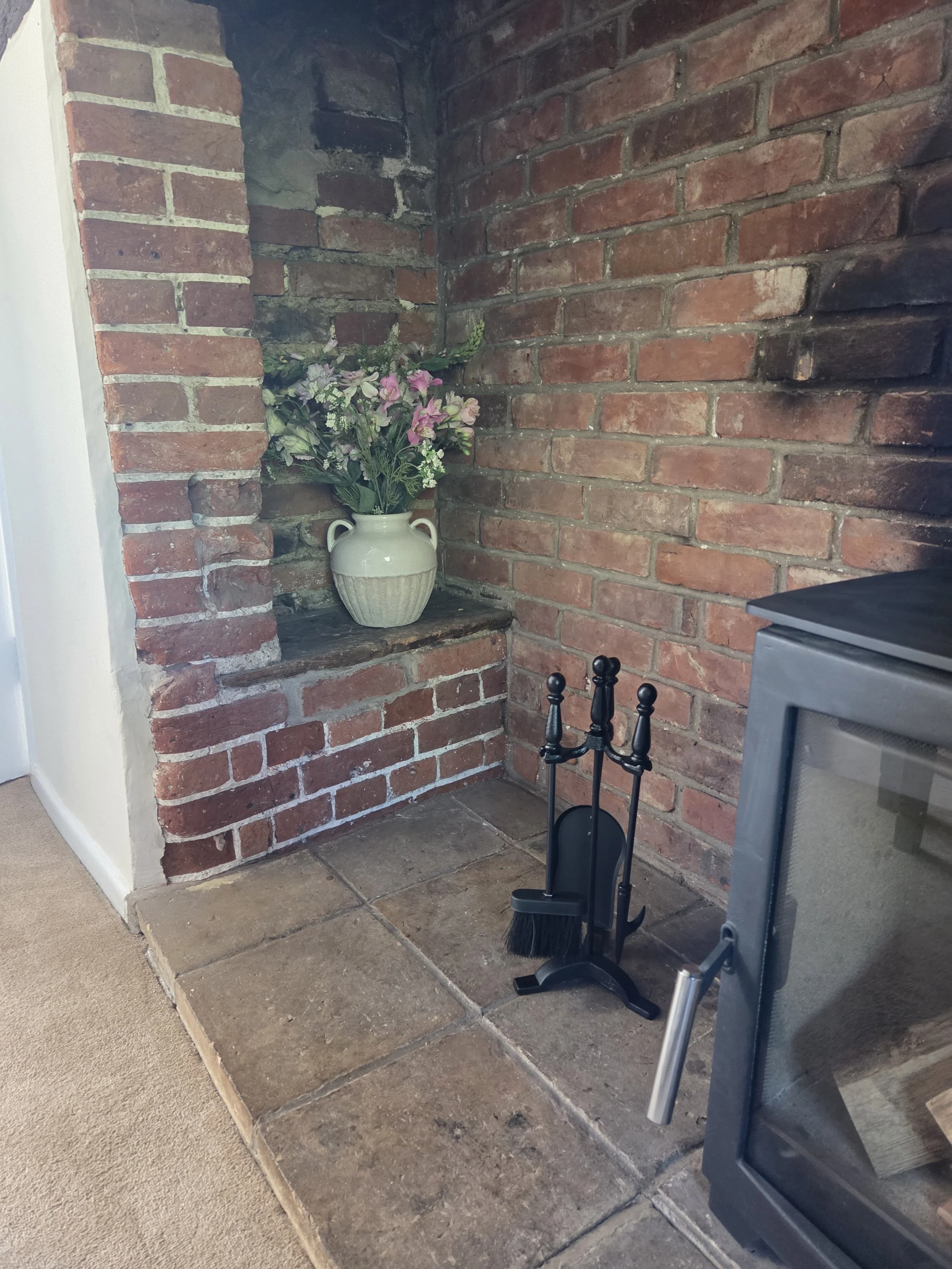 A corner with a brick wall and a white vase with pink and purple flowers on a shelf. There are fireplace tools in front of the wall, and a stove to the right. The floor is a mix of tiles and carpet.