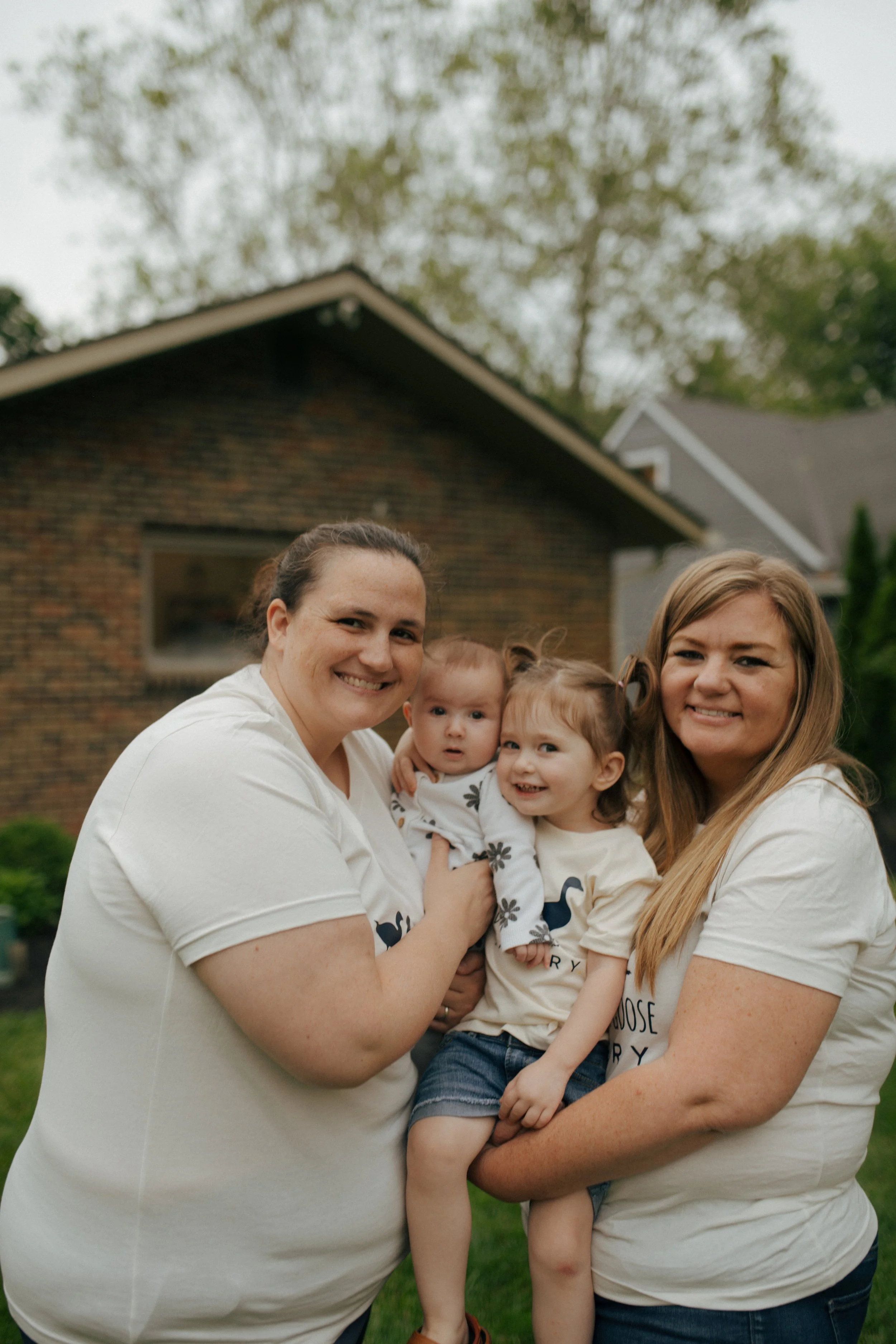 Owner of Duck Duck Goose Bakery, Cara holding and hugging her kids and wife in a grassy outdoor area with trees and a house in the background.