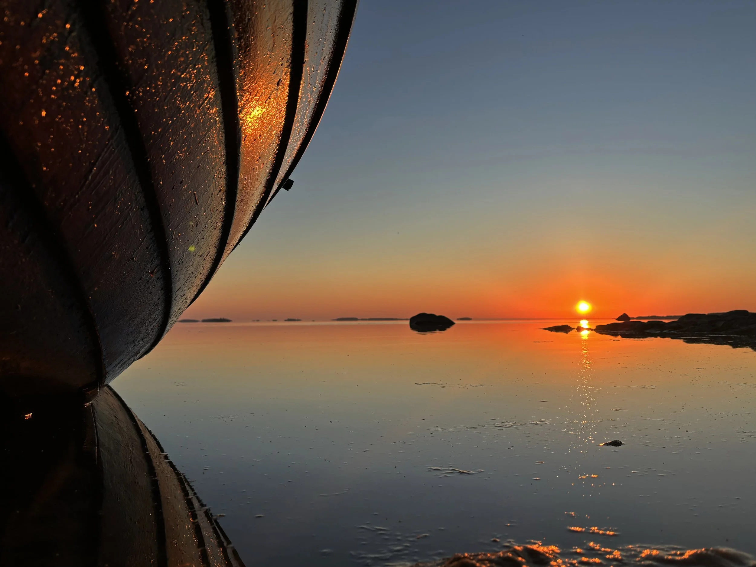 Close-up view of the front of a boat with water droplets, during sunset with the sun near the horizon over calm water and rocks in the background.