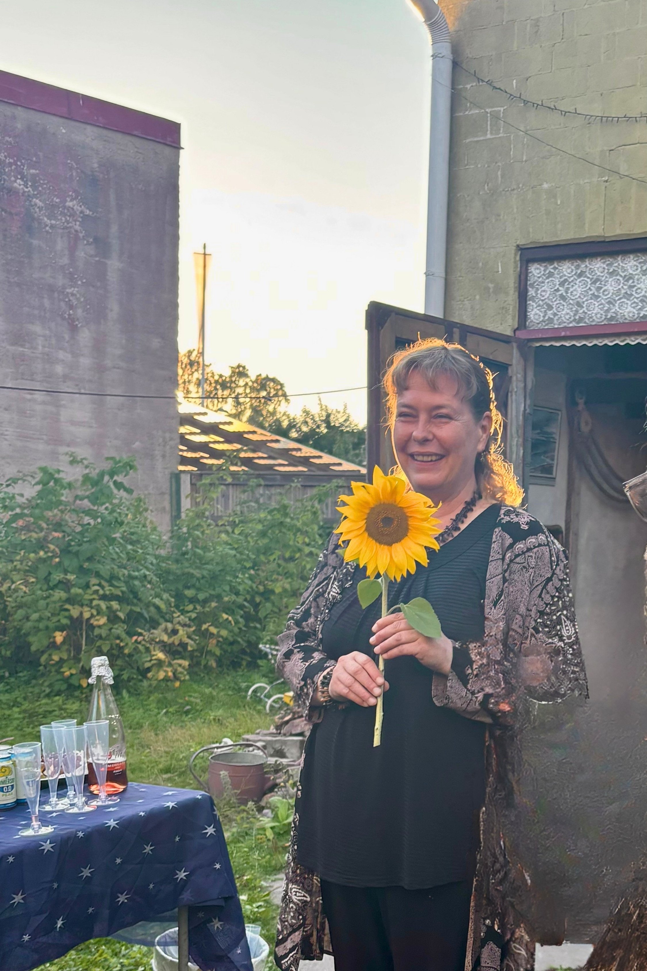 A smiling woman holding a large sunflower in the lush green courtyard of Galleria Kissaniitty in Reposaari, Pori. On the left, a serving table with sparkling wine glasses is visible under the bright afternoon light.